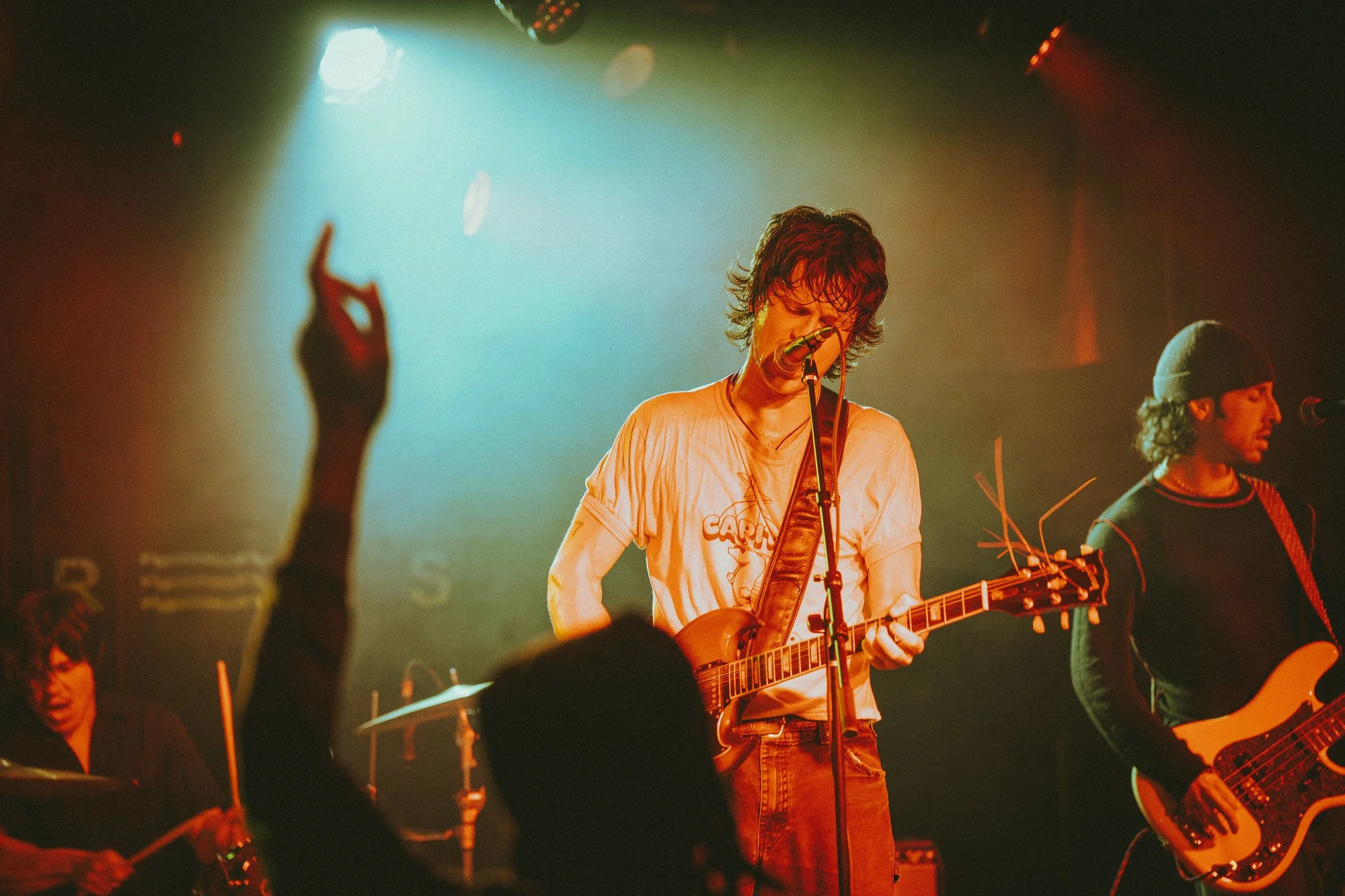Musicians performing on stage with one playing an electric guitar and singing into a microphone, another playing a bass guitar, in a dimly lit venue with audience in foreground.