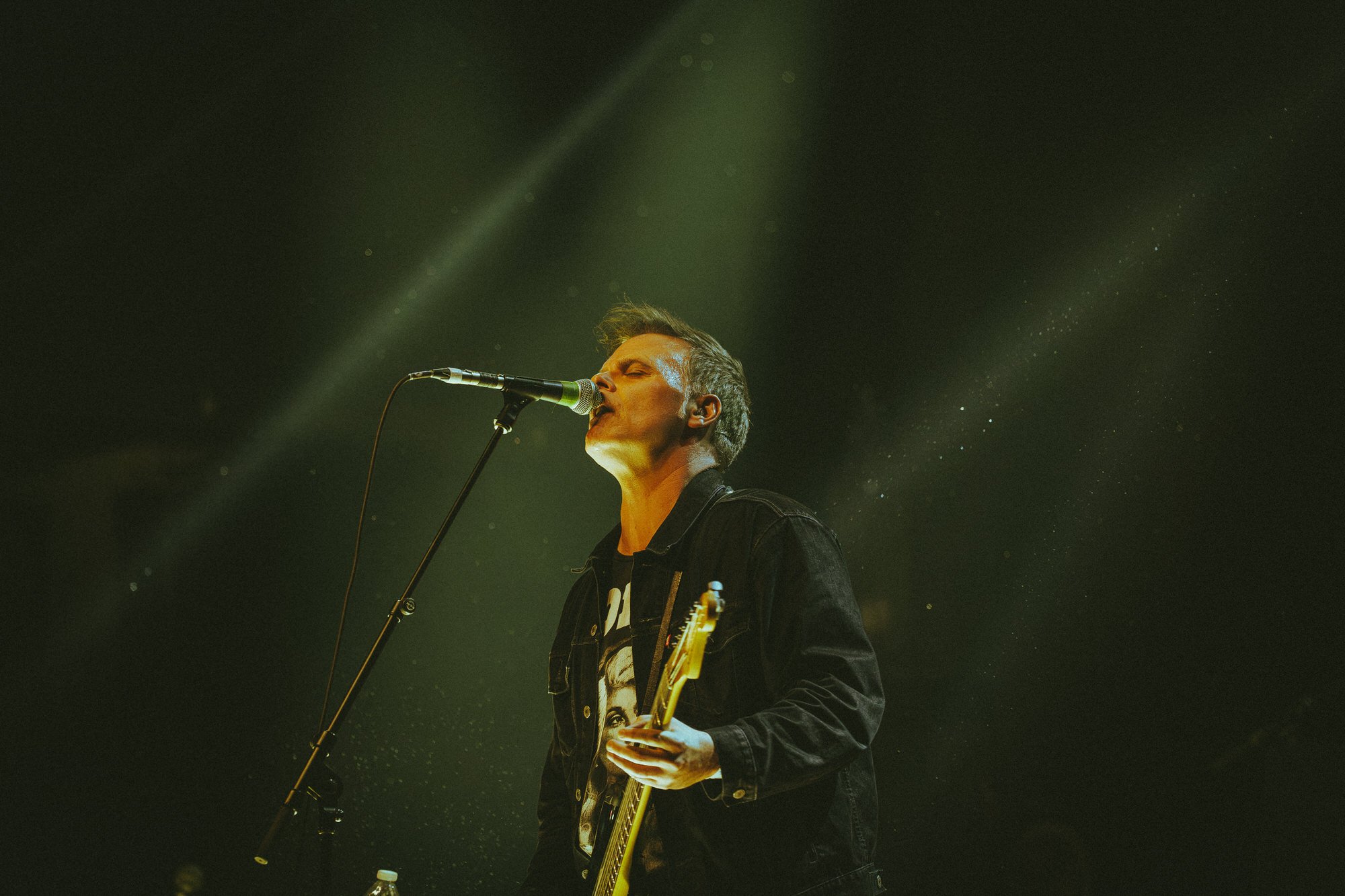 Male musician singing into a microphone while playing an electric guitar on stage with green and yellow lighting.