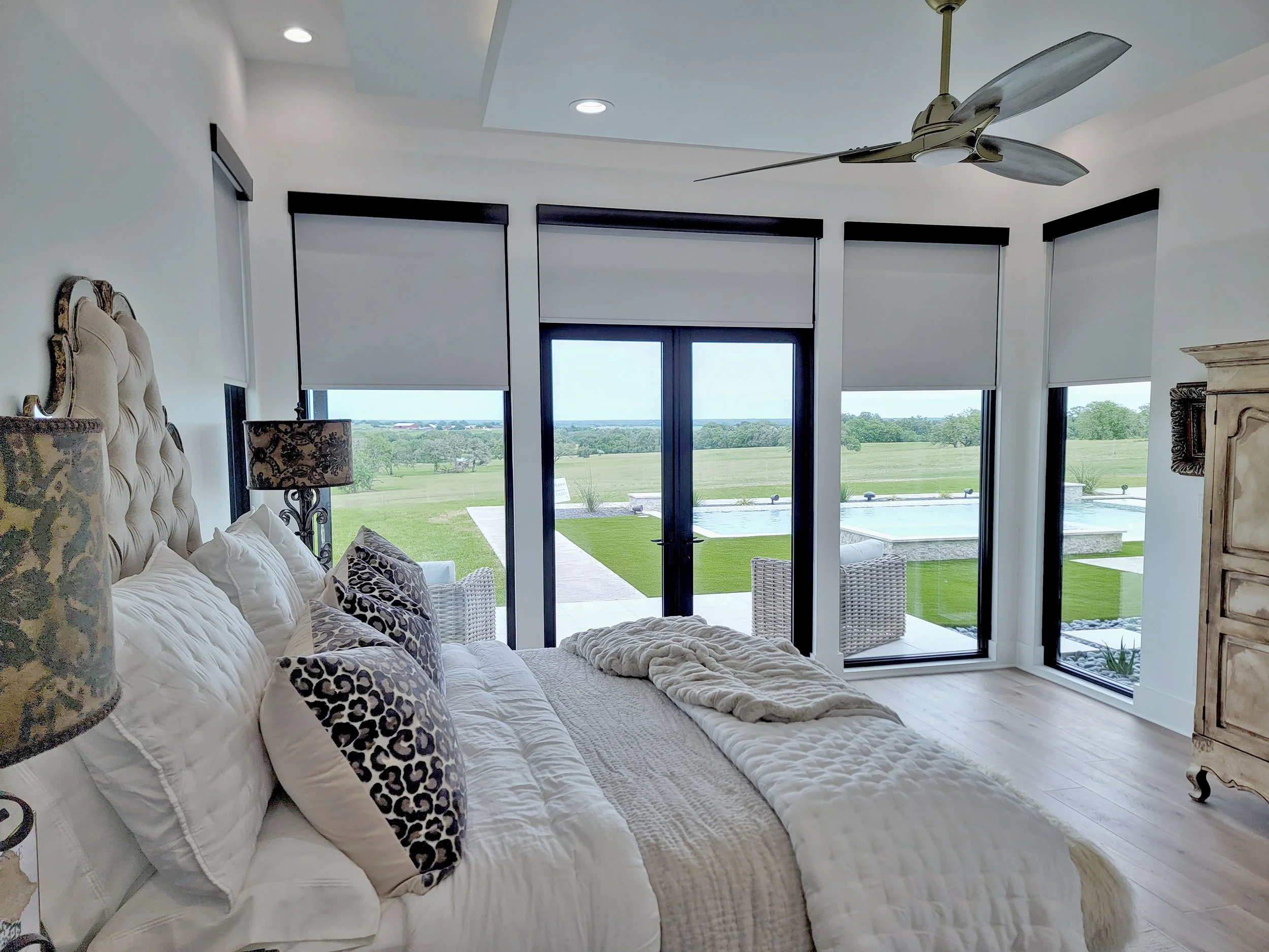 Custom roller shades installed on large bedroom windows and glass doors overlooking an outdoor pool and landscape.