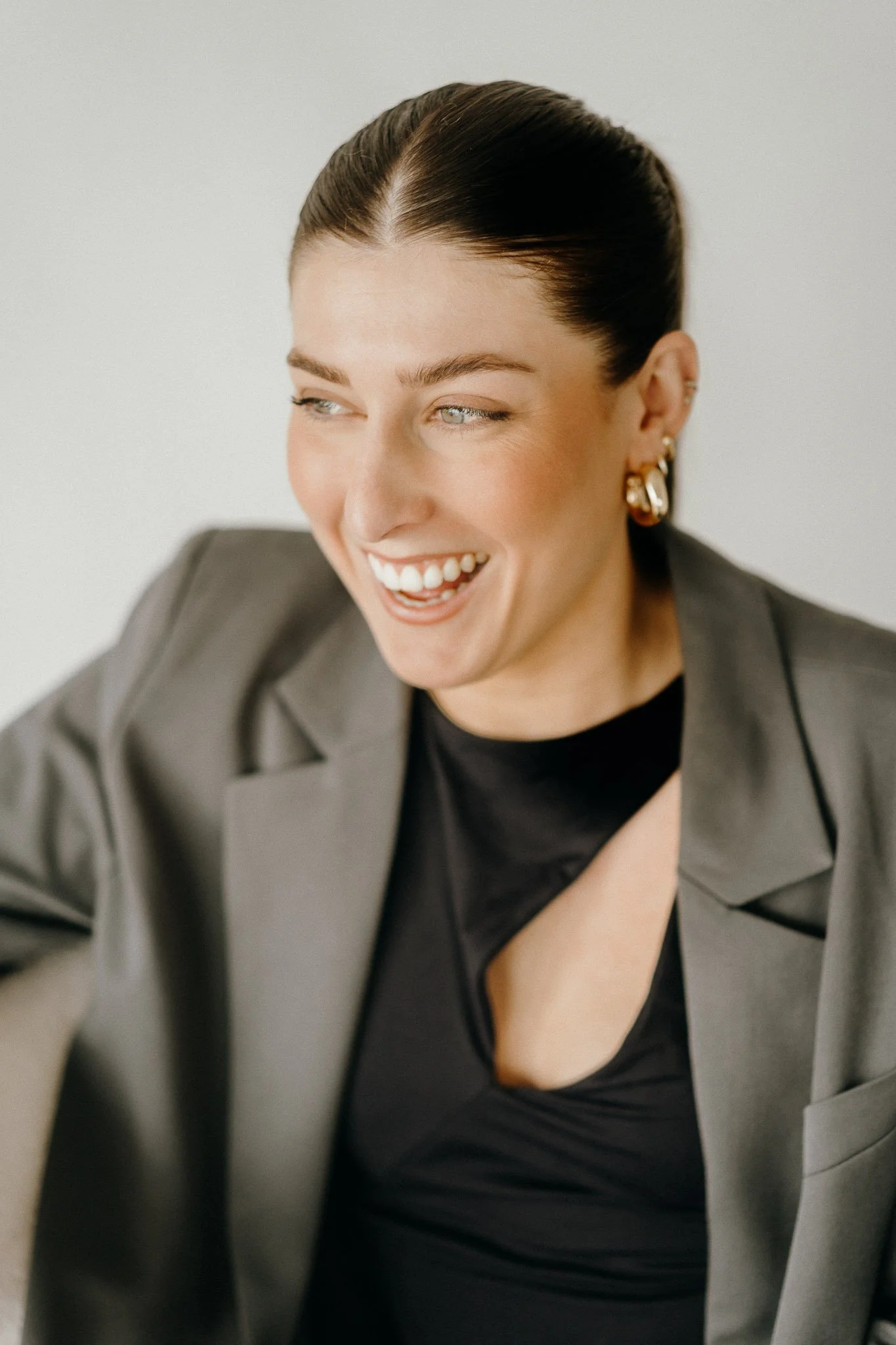 Smiling woman in a black top and gray blazer, wearing gold hoop earrings, with hair tied back.