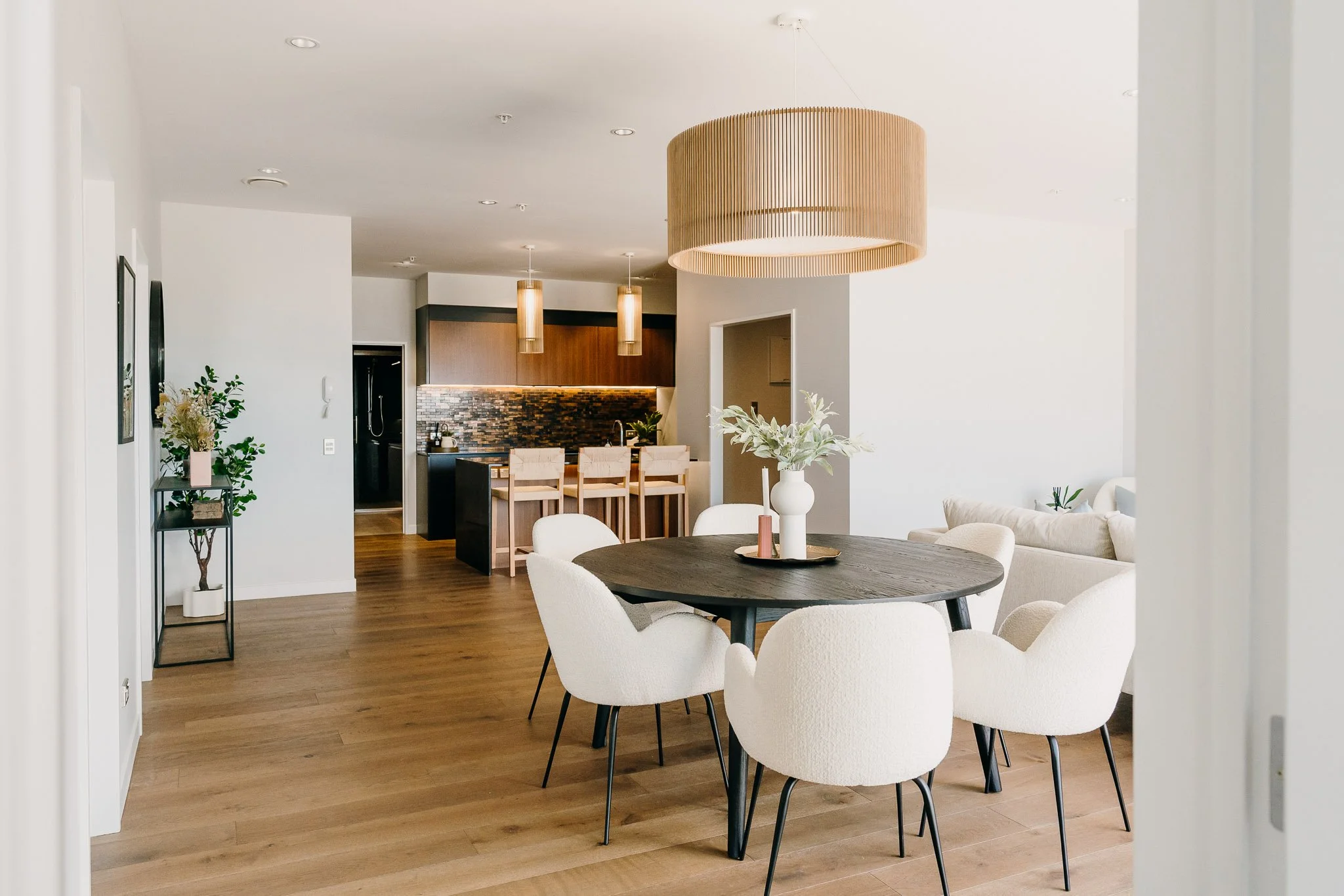 Modern open-plan dining and kitchen area with black round table, white chairs, large pendant light, wooden flooring, kitchen island with bar stools, and contemporary decor.