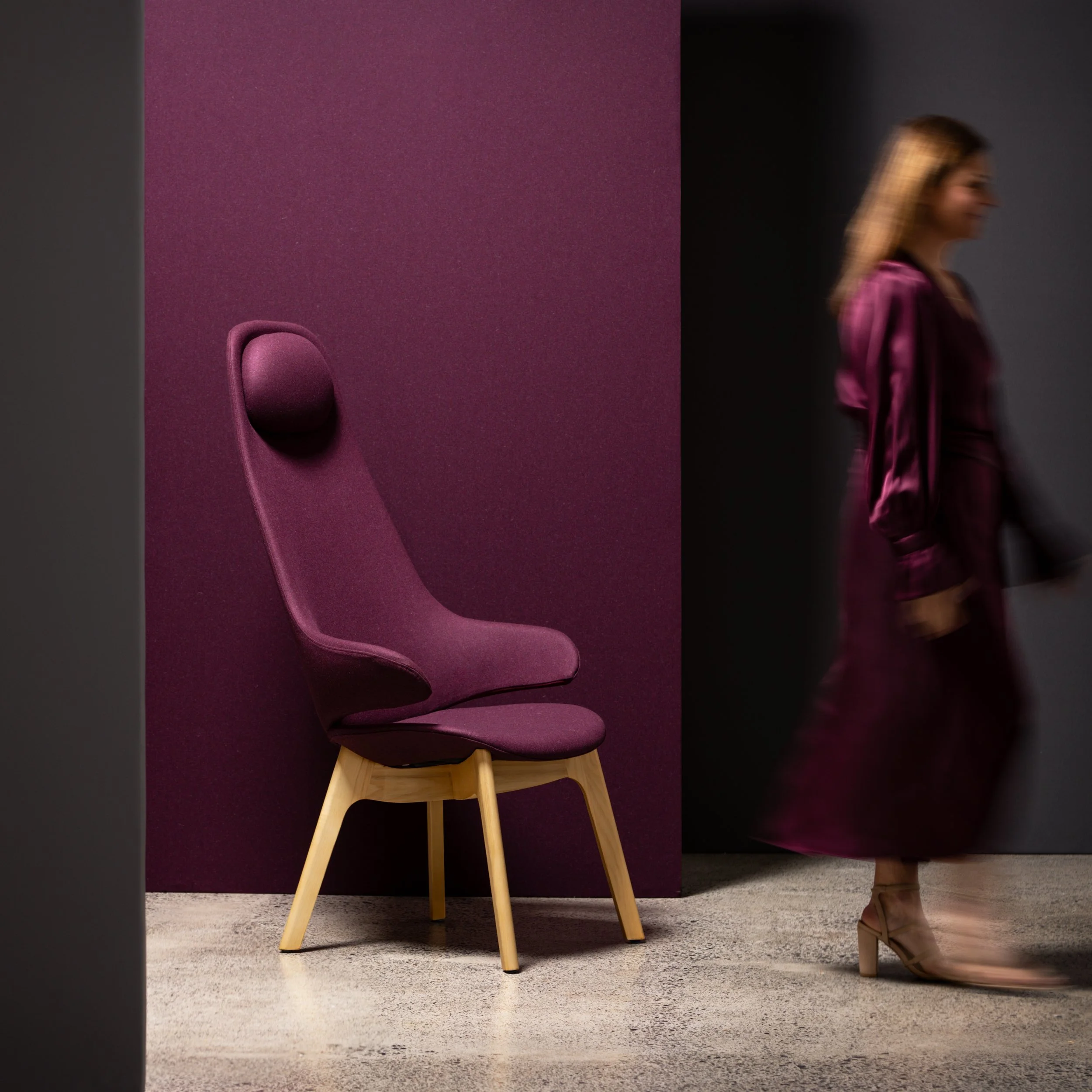 Curated studio furniture photograph featuring a burgundy fabric upholstered chair and matching fabric backdrop, with a model in complementary tones walking through the frame; styled with grey accents for a commercial shoot. 