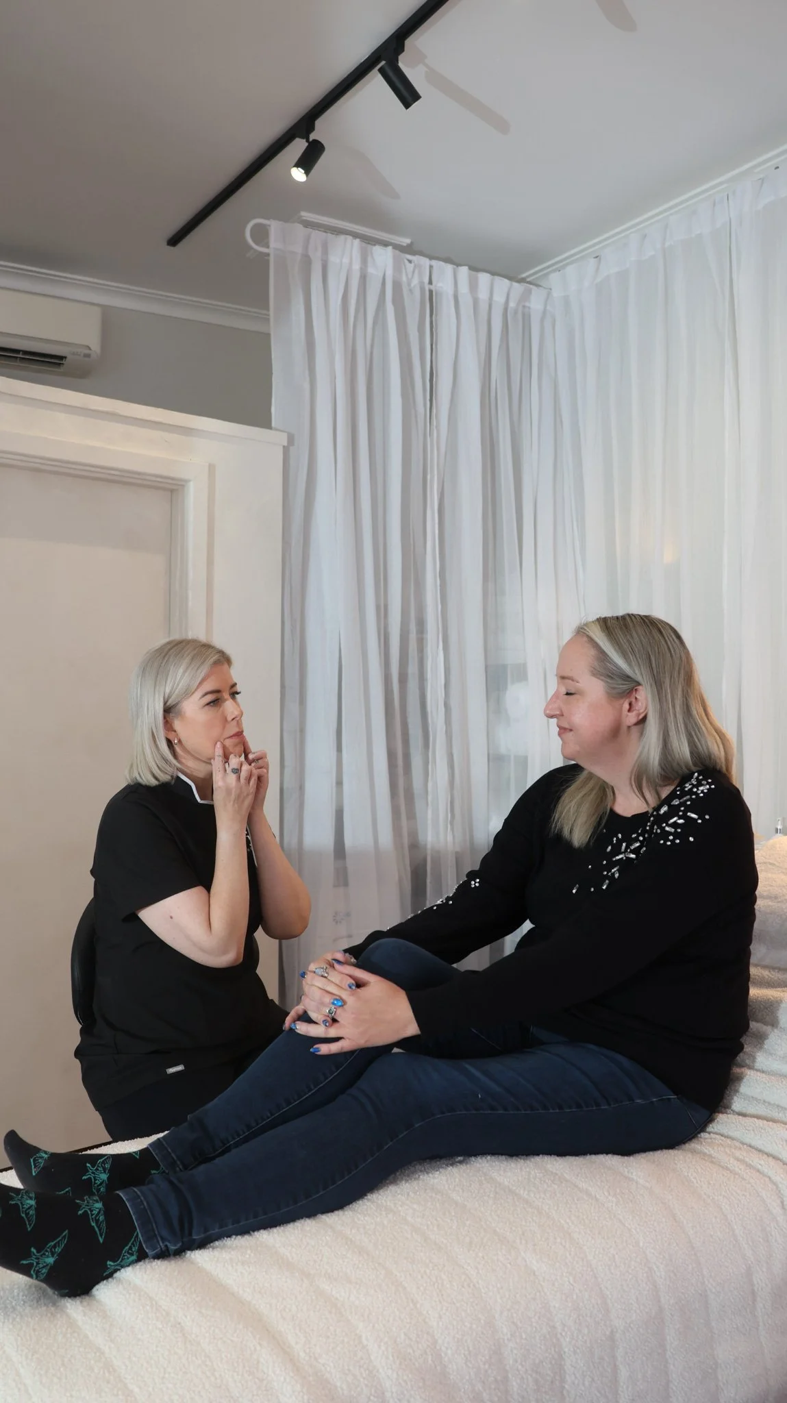 Two women sit close together on a white bed in an aesthetic treatment room with white curtains, engaging in a conversation. One woman has platinum blonde hair and is wearing a black top, appearing attentive.