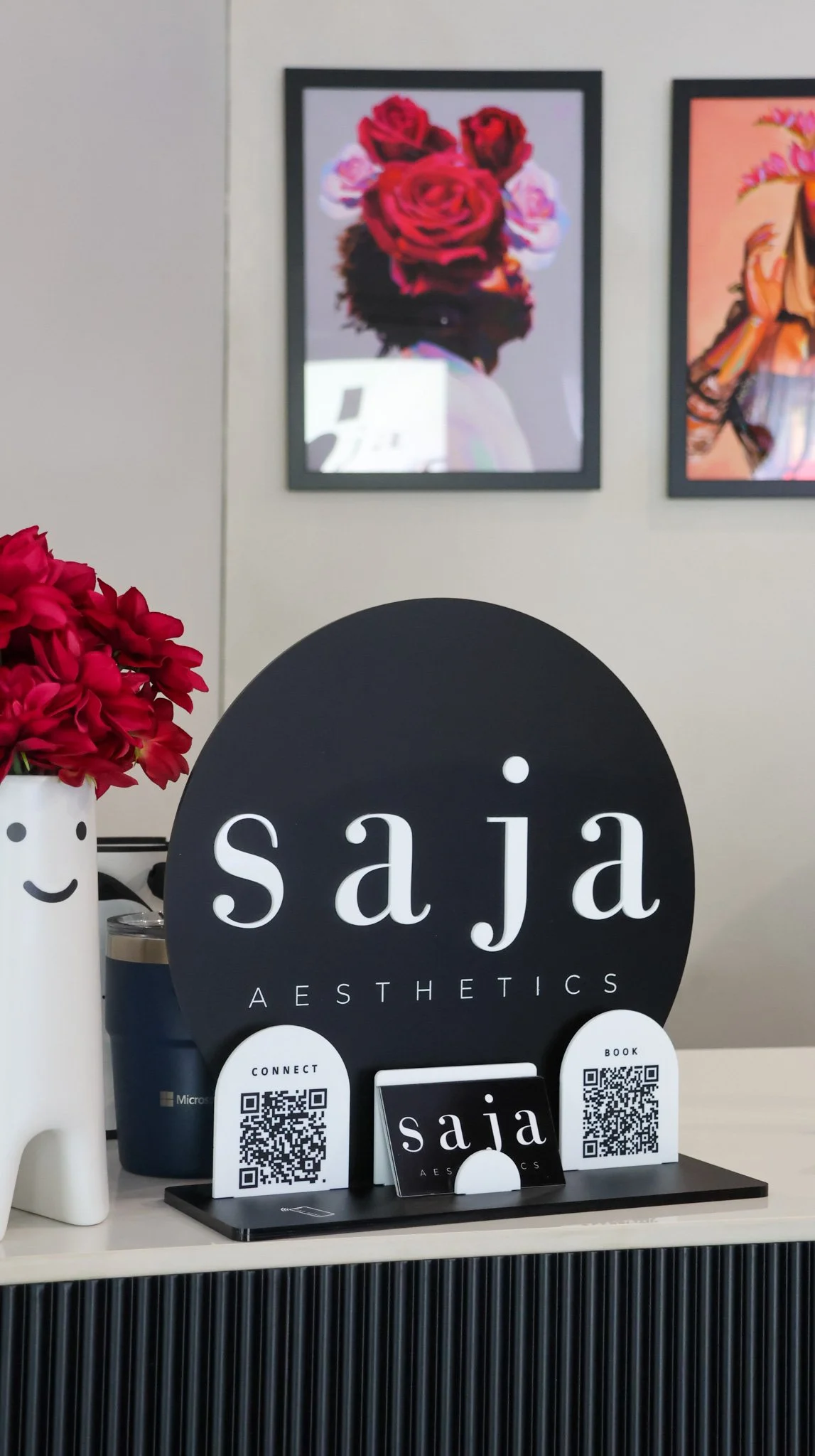 Display stand for Saja Aesthetics with QR codes and a card featuring the logo. Behind it, a vase with red flowers, and a framed artwork of a woman with a floral headpiece is mounted on the wall.