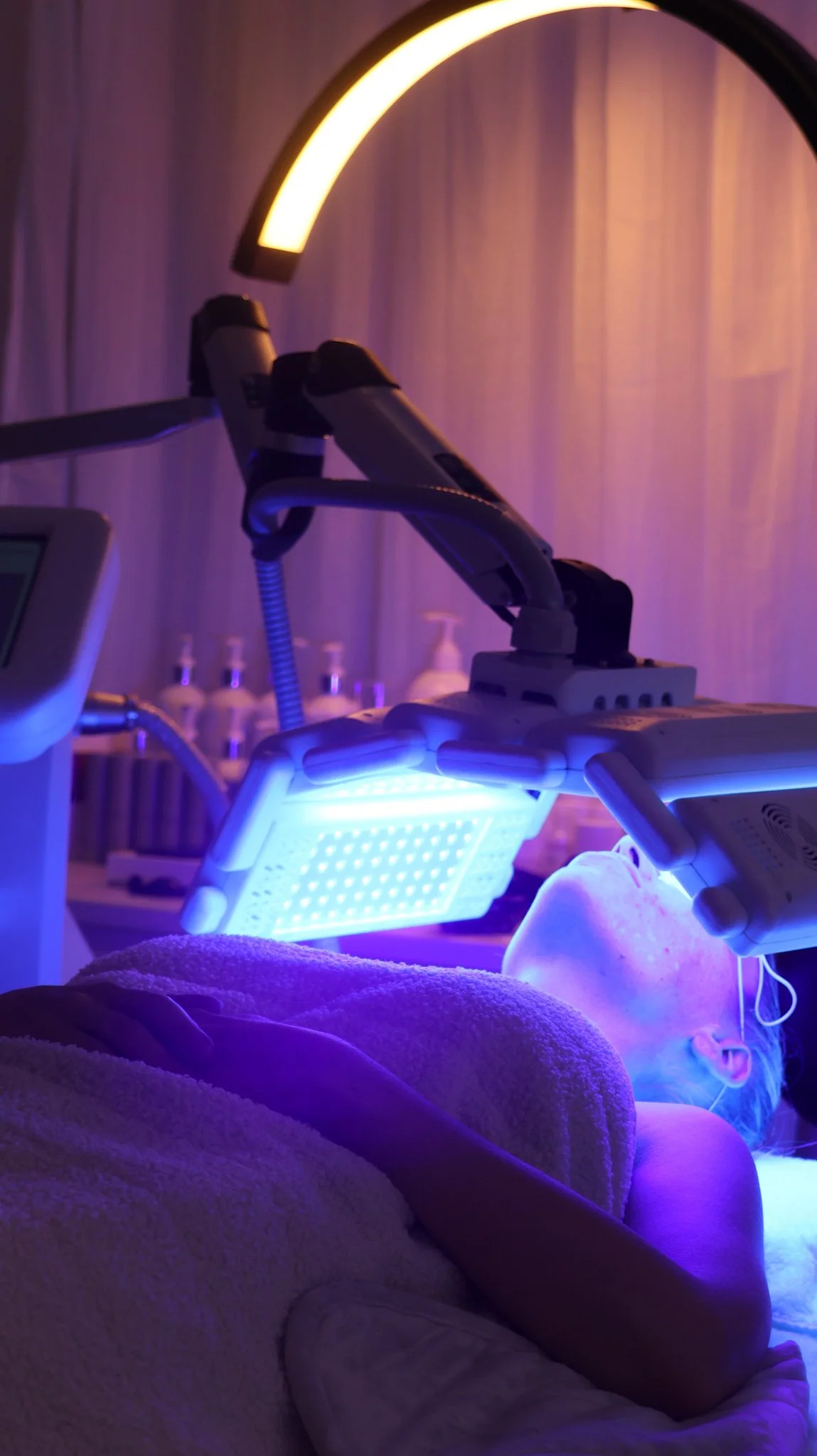 A person receiving a facial treatment under blue LED light therapy, with a facial mask applied. The scene is in a spa or medical clinic with medical equipment and bottles in the background, illuminated by blue and purple lighting.