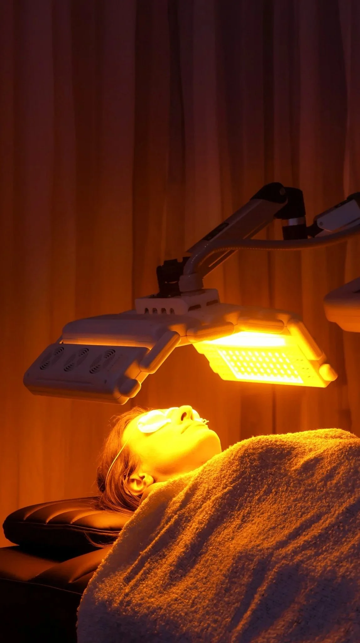 A woman lying down with her eyes closed under a yellow heat lamp, wearing protective eye goggles, in a room with wooden walls.