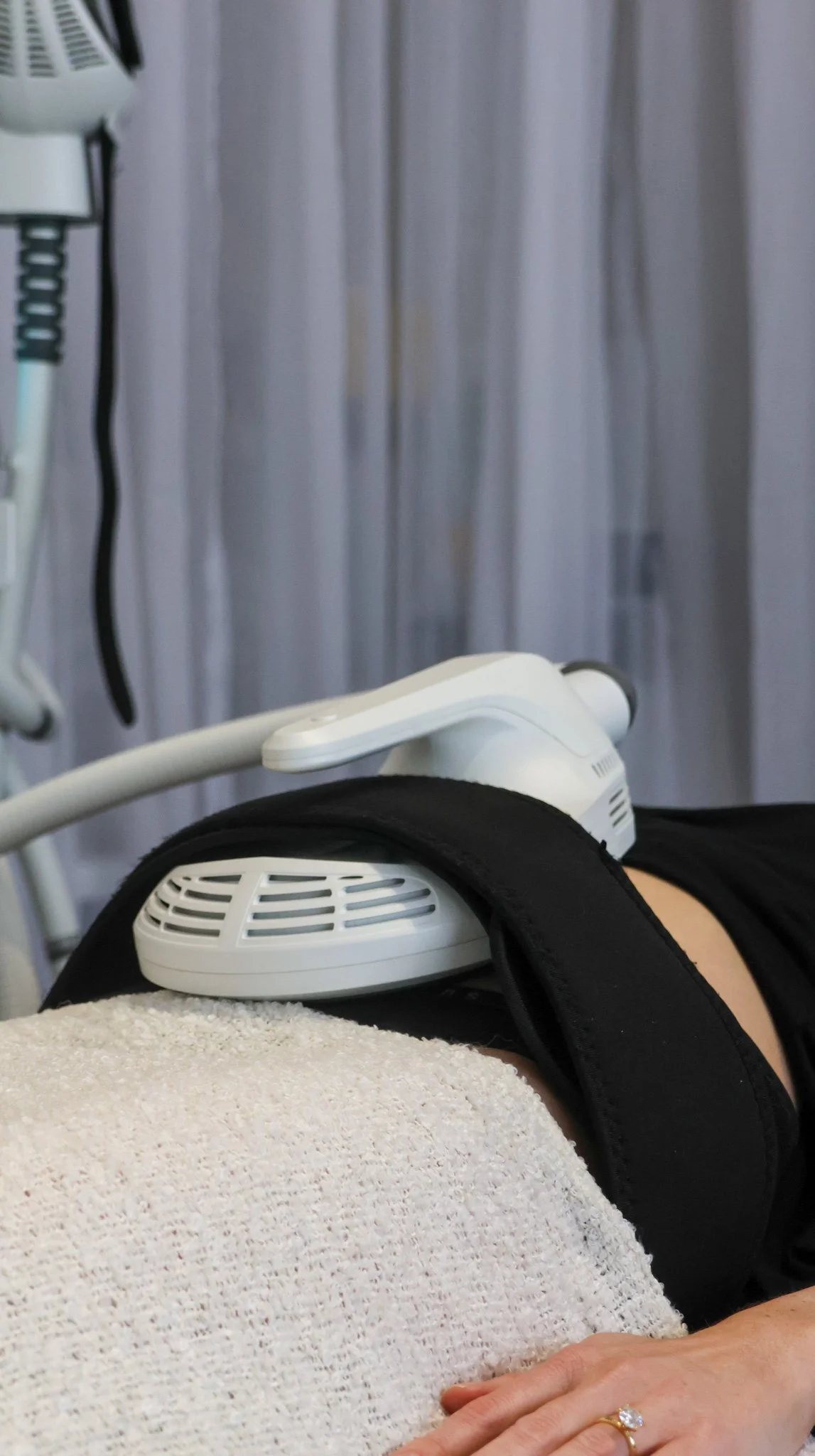 Person receiving a medical or cosmetic fat freezing treatment with a device on their abdomen, in a clinical setting with a curtain in the background.
