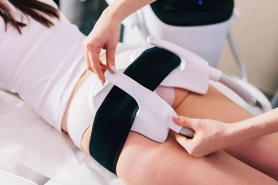 Person undergoing a body contouring procedure using a device on their thigh, with a healthcare professional adjusting the equipment.