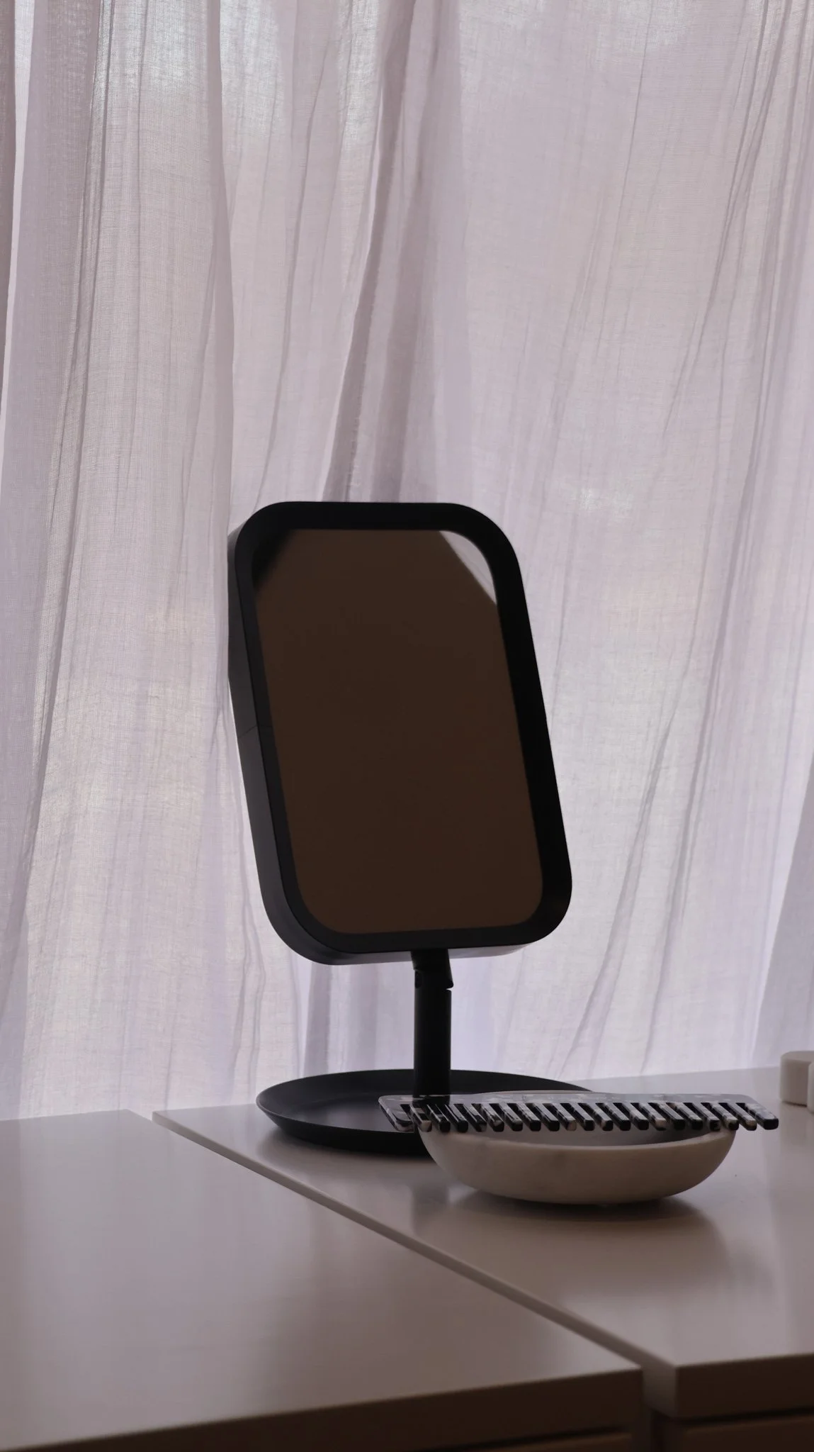 A black-framed mirror on a stand, placed on a white table next to a bowl with a black grate and a white rectangular object, with white curtains in the background.