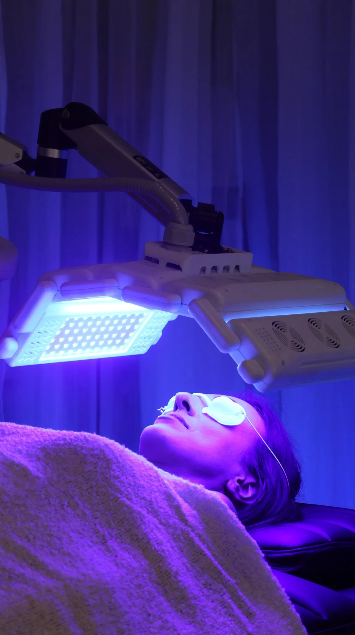 A woman undergoing a laser or LED skin treatment in a medical or spa setting, lying on a bed with her eyes covered, under a purple-blue medical light