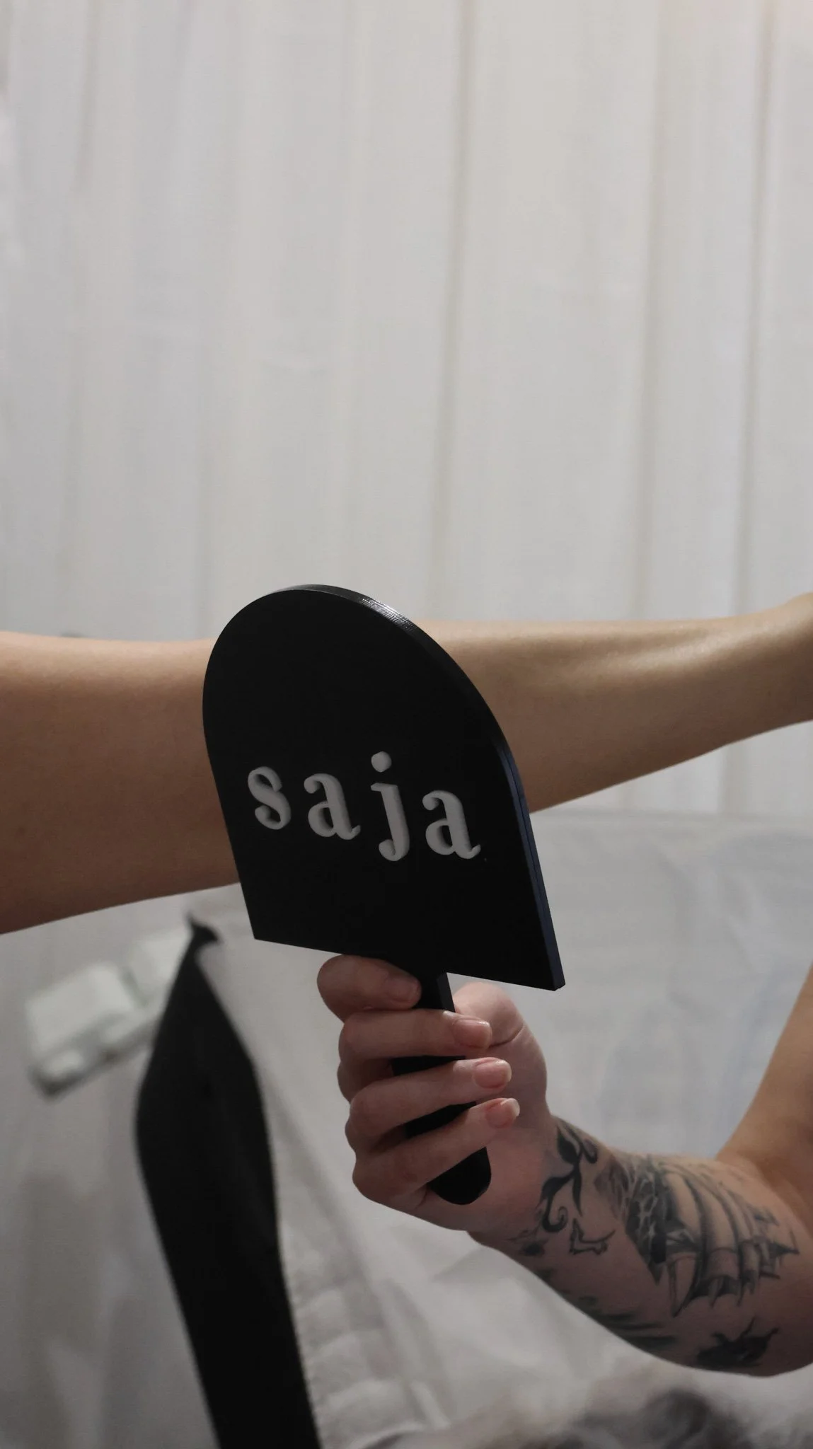 Person with tattooed arm holding a sign that says 'saja' in front of a person with a bare shoulder, in a room with white paneled wall.
