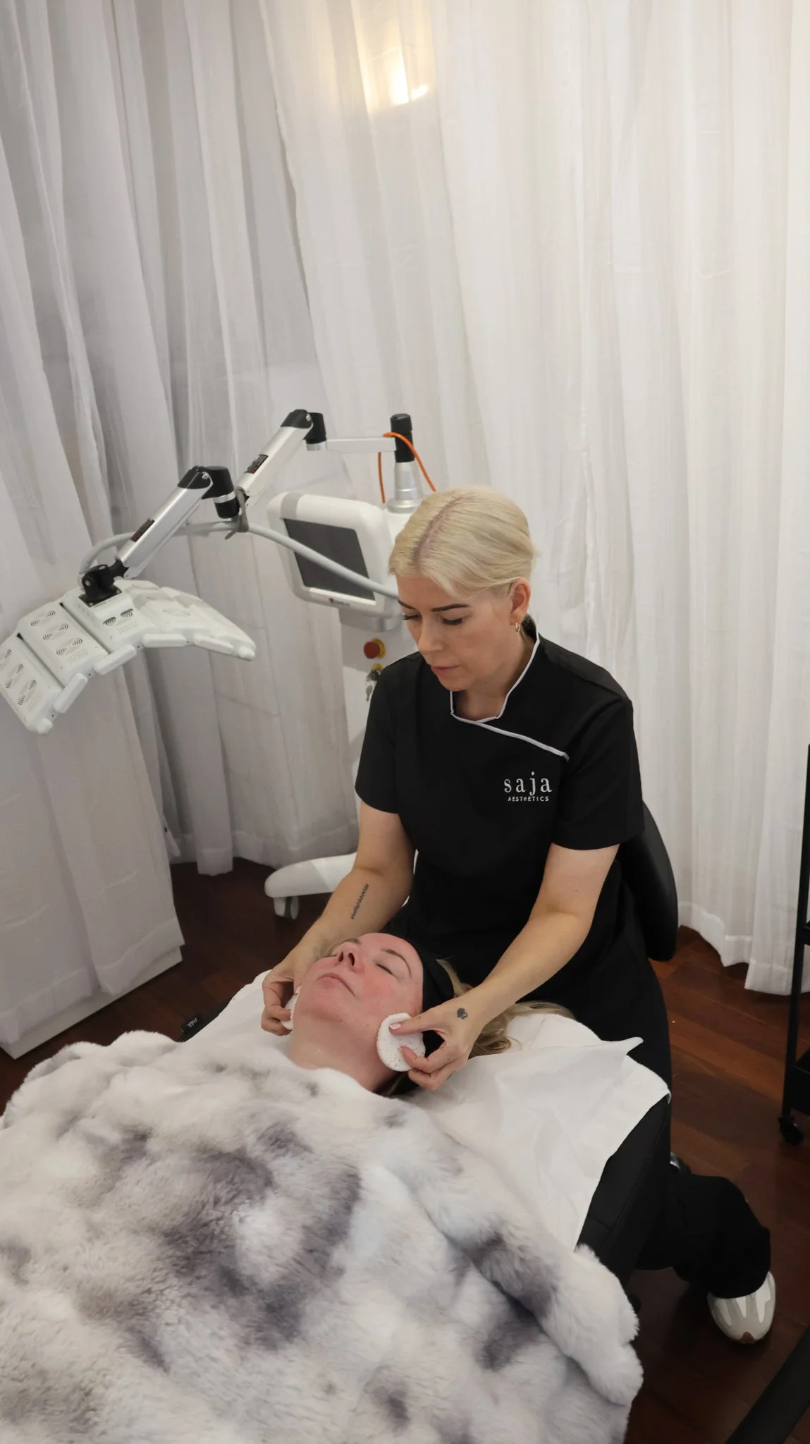 A woman receiving a facial treatment from a professional aesthetician in a treatment room. The woman is lying on a bed covered with a white and gray blanket, while the aesthetician uses a cloth to clean her face. There are medical equipment and a monitor in the background.