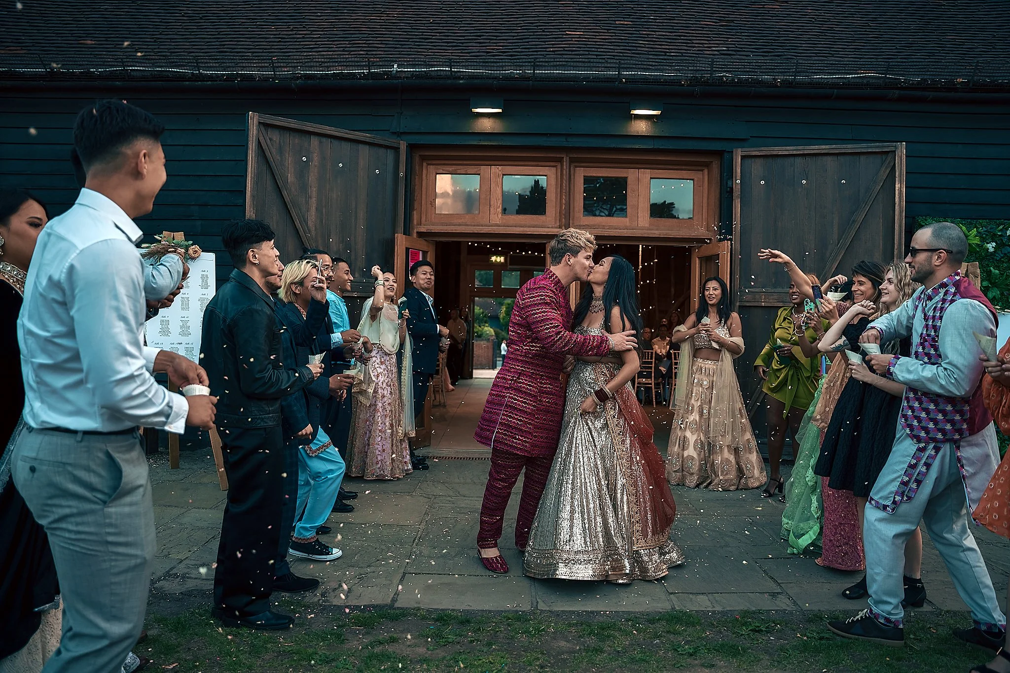 A wedding celebration outside a barn with a couple sharing a kiss in the center, surrounded by friends and family throwing confetti.