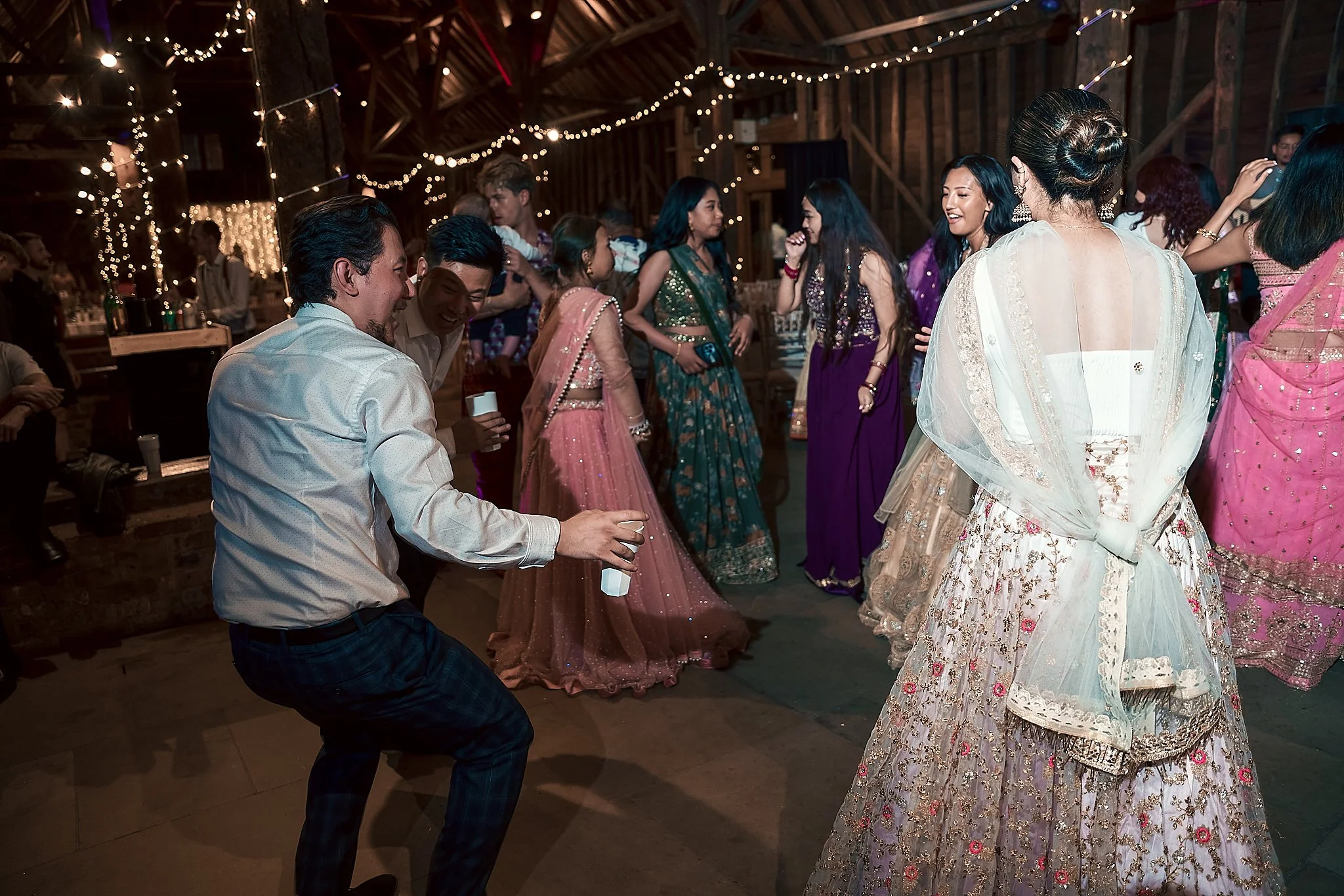 People dancing and celebrating at a festive event with string lights hanging from the ceiling, many women in colorful traditional dresses and men enjoying the lively atmosphere inside a rustic barn.