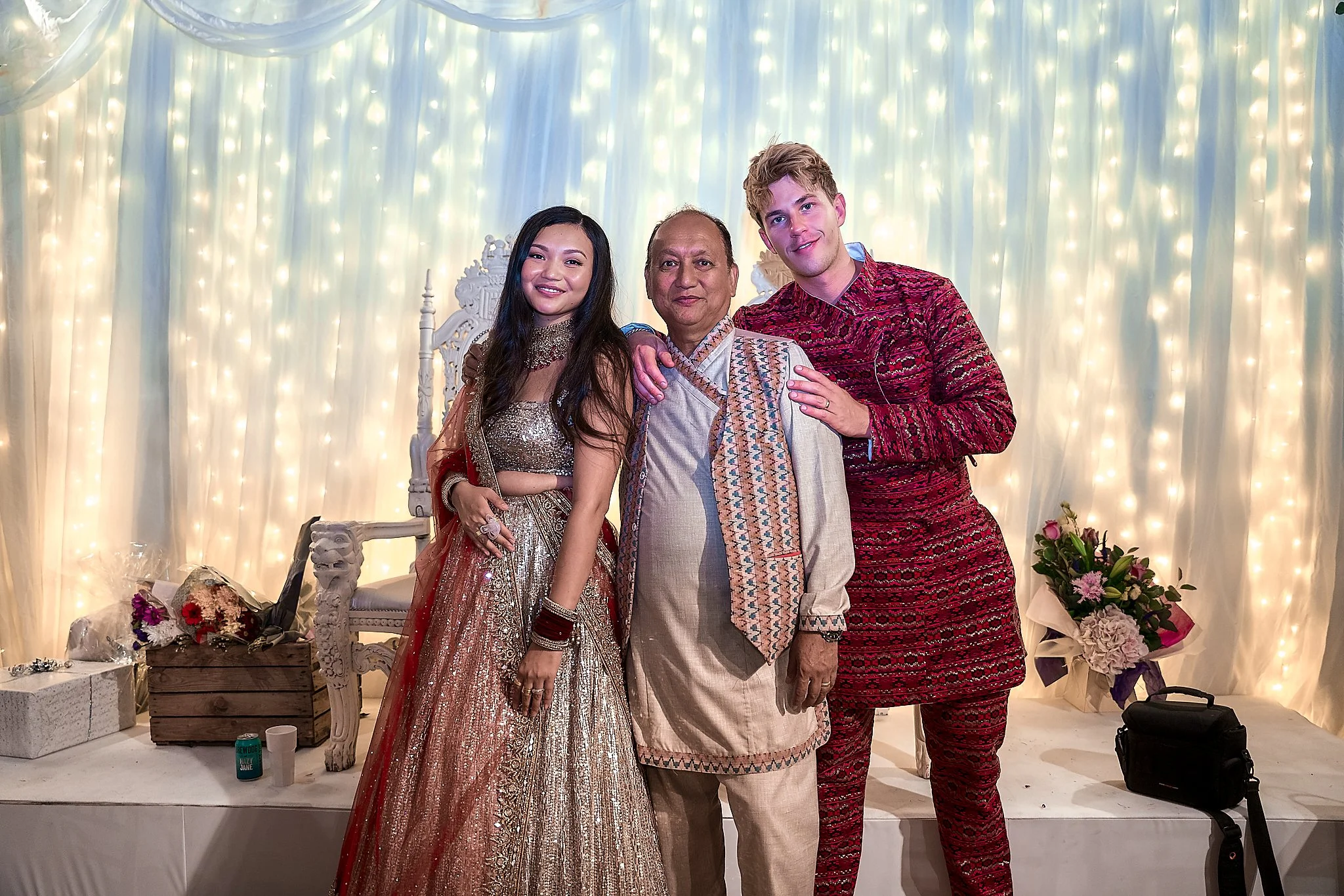 Three people in traditional Indian attire posing together in front of a decorated backdrop with white curtain and string lights, with flowers and gifts on the side.