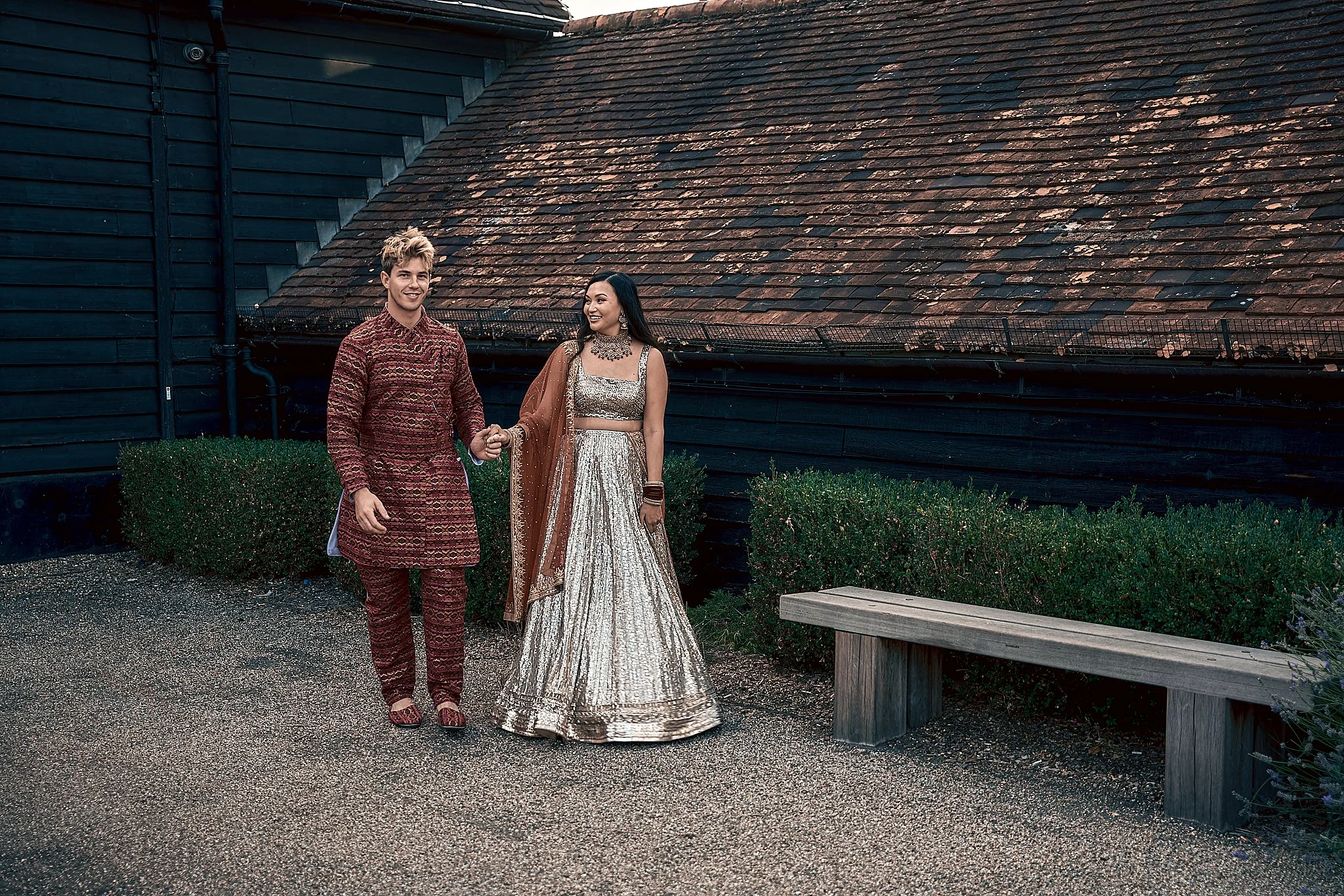 A man and woman dressed in traditional Indian attire, walking hand in hand outdoors near a dark wooden building with a sloped roof. The woman is wearing a silver and gold lehenga with jewelry and a dupatta, and the man is in a matching maroon kurta a