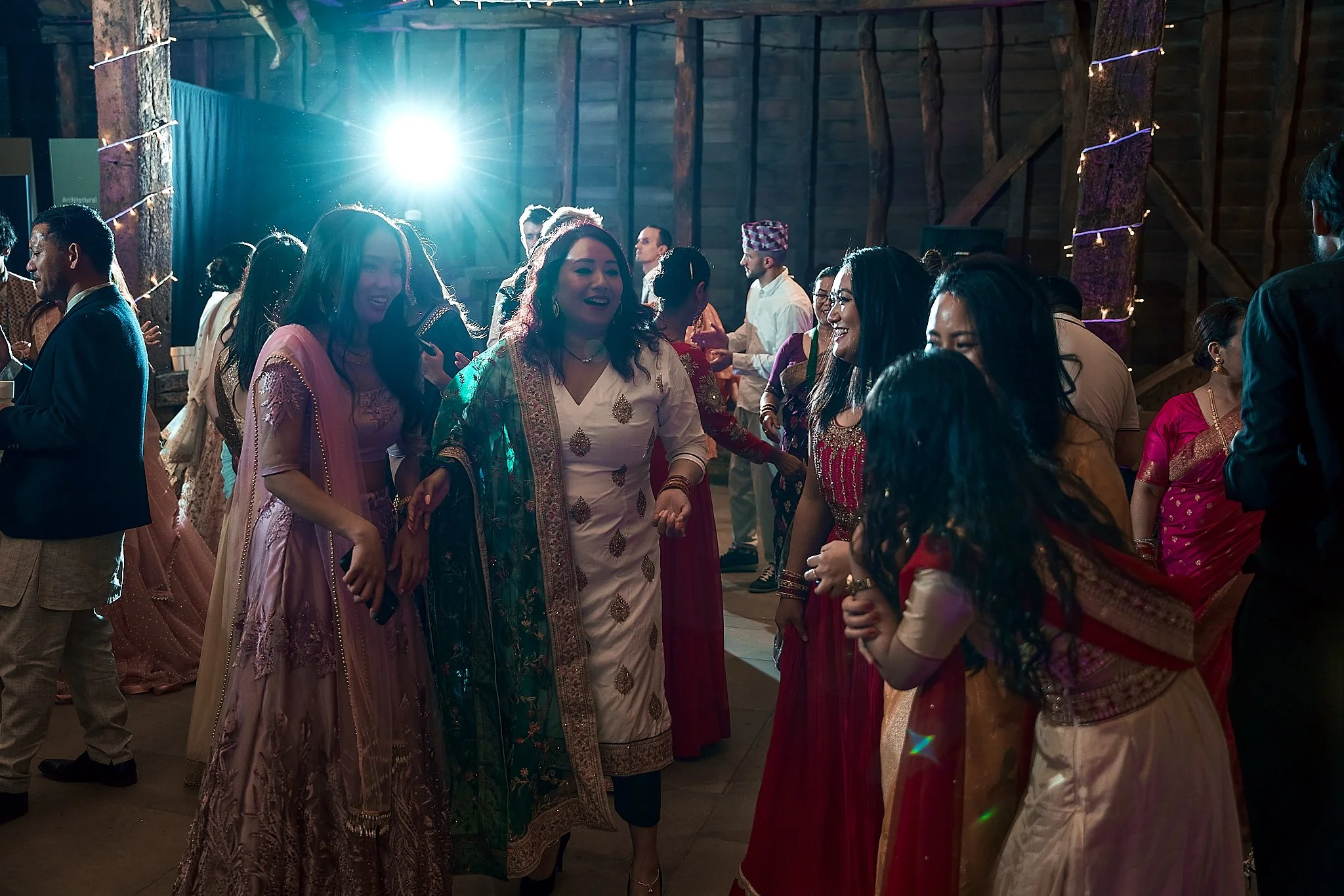 Group of people dancing and socializing at a celebration or party in a rustic wooden venue, dressed in traditional South Asian attire, with a bright light in the background.