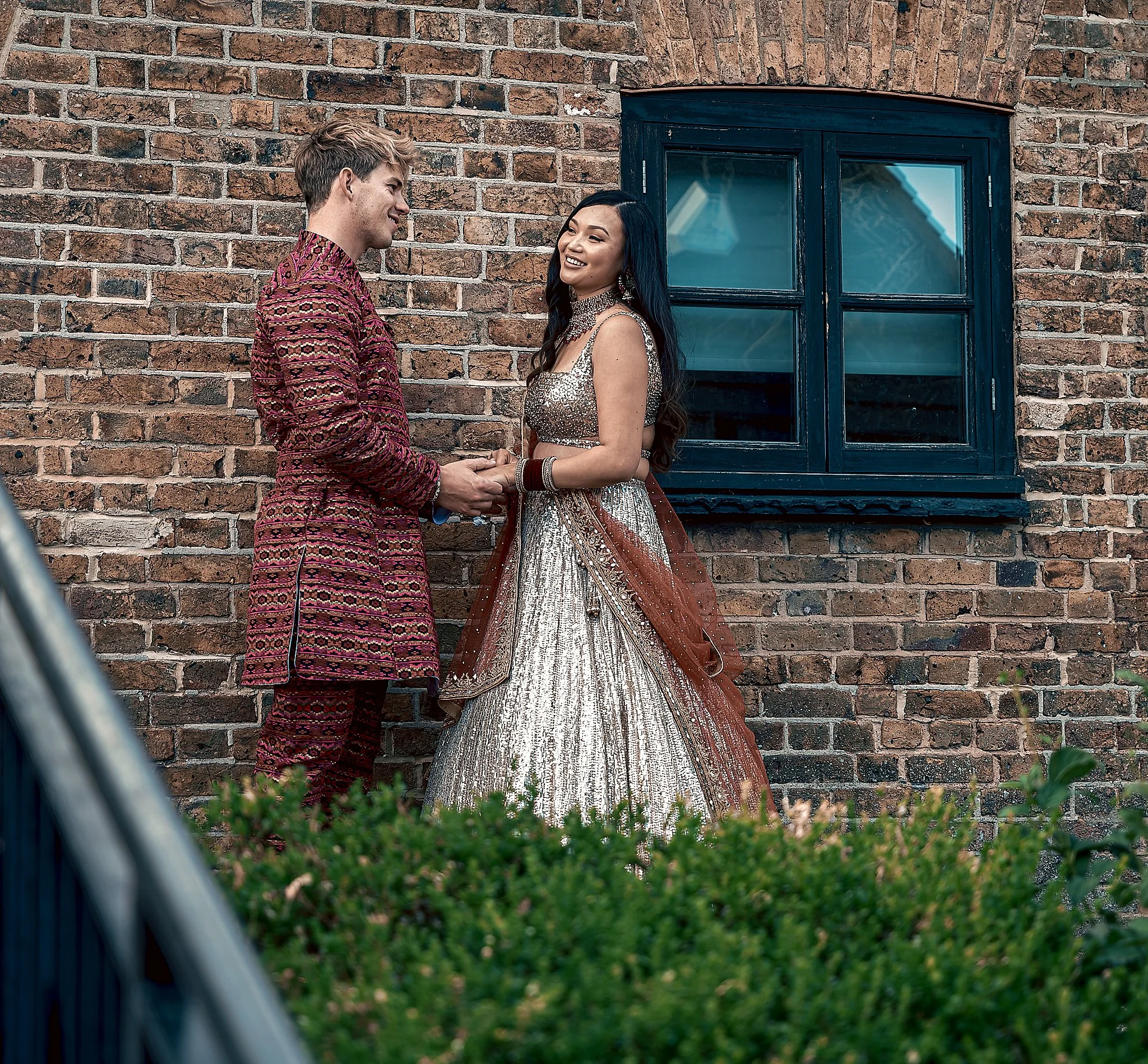 A couple dressed in traditional Indian wedding attire standing outdoors against a brick wall and window, holding hands and smiling at each other.