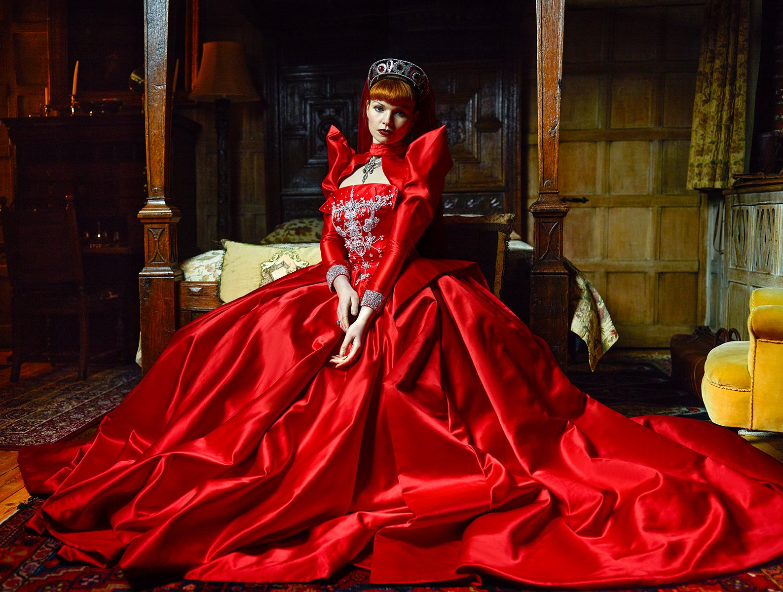A woman in a luxurious red gown sitting on a vintage bed in a richly decorated room.