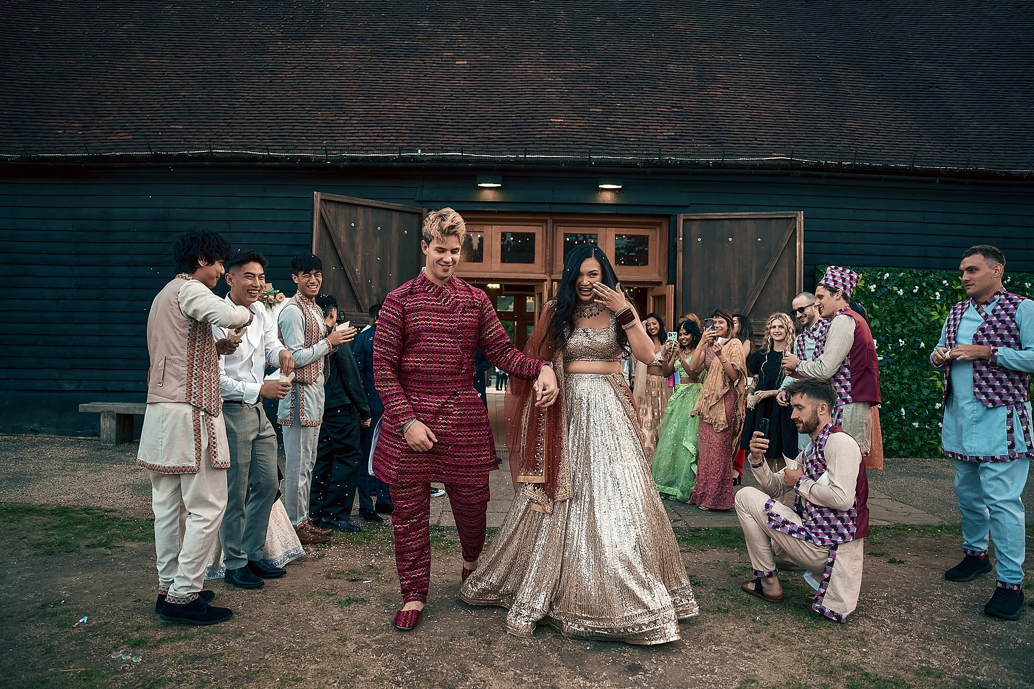 A couple in traditional Indian attire walking at an outdoor wedding celebration, with guests watching and taking photos in the background.
