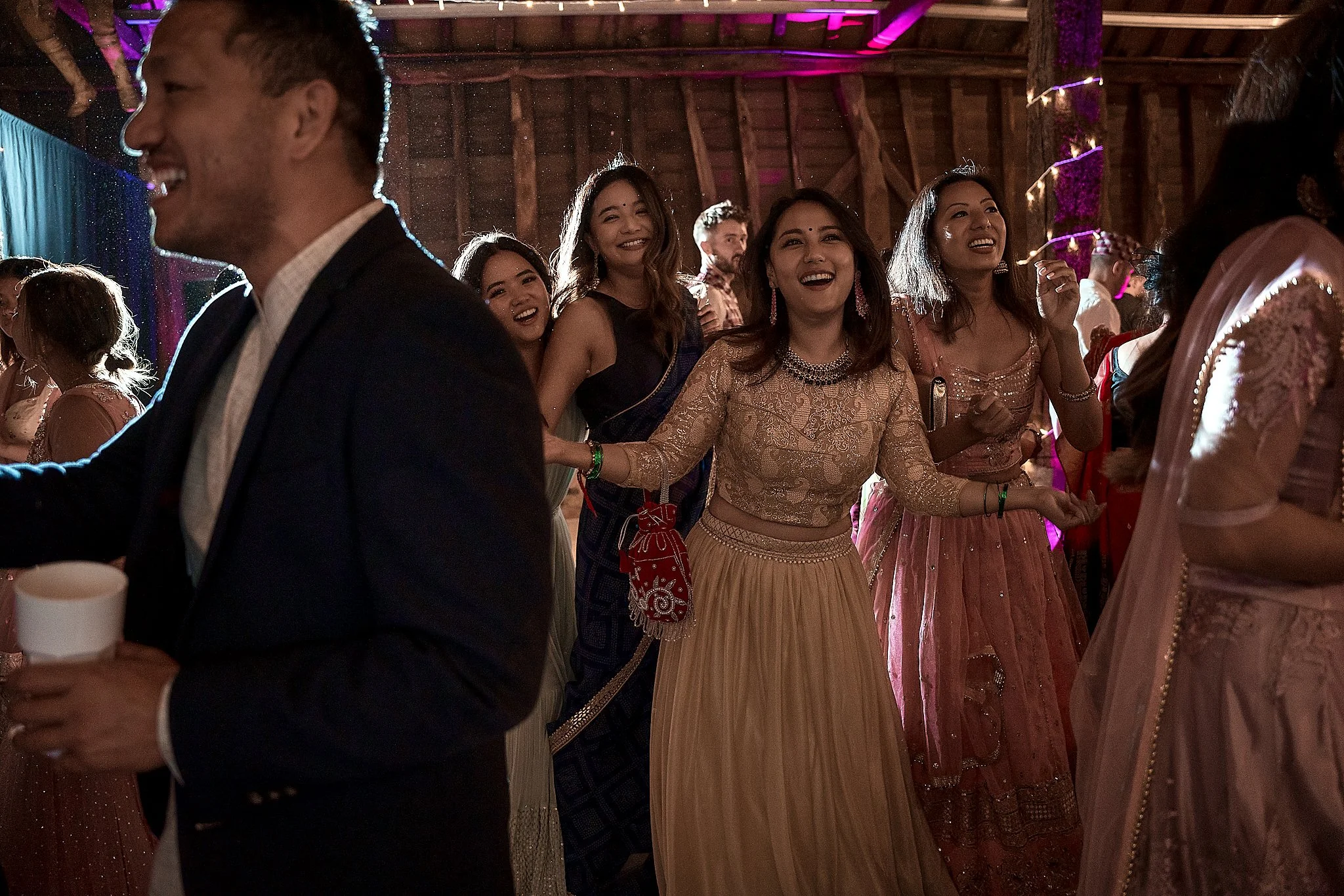 People dancing and enjoying a celebration in a rustic venue with wooden beams, some wearing traditional Indian attire.