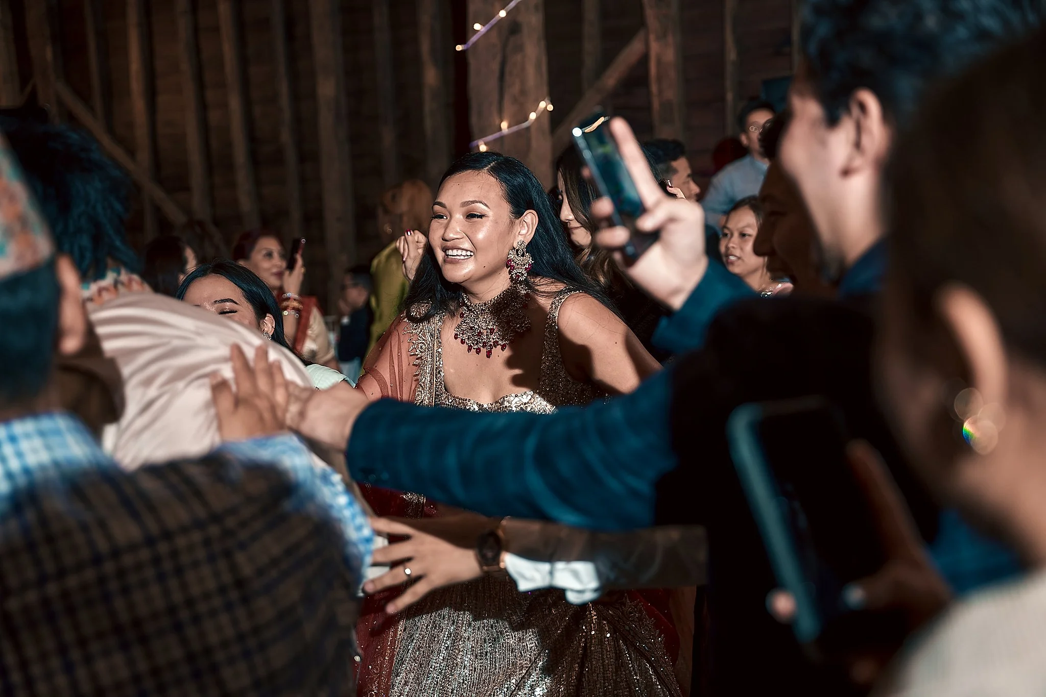 A woman in traditional Indian attire smiling and dancing at a celebration, surrounded by other people taking photos and enjoying the event.