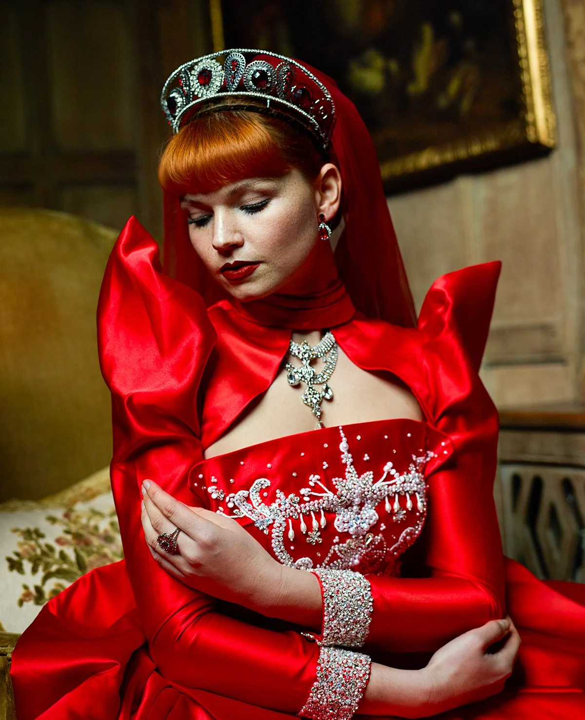 A woman dressed in an ornate red gown with silver embellishments, wearing a tiara, earrings, and a necklace, with her eyes closed and arms crossed over her chest, sitting in a richly decorated room.