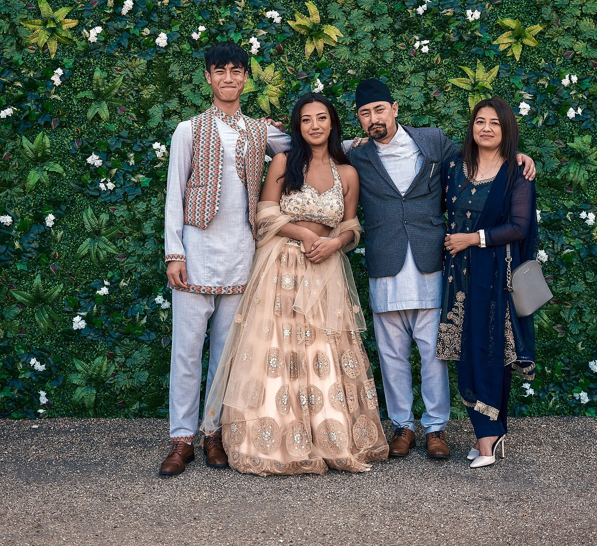 Group of four people standing in front of a green leafy wall decorated with white flowers, dressed in traditional South Asian attire for a celebration.