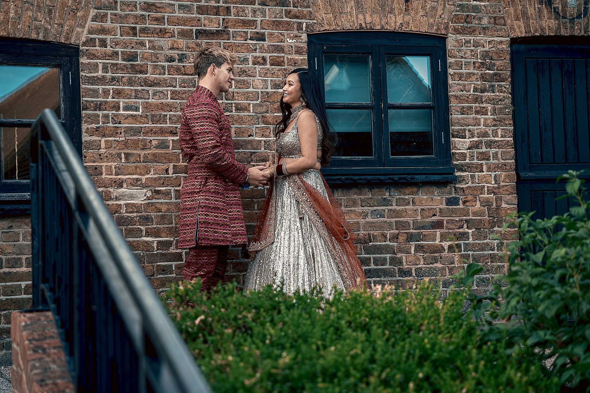 A couple dressed in traditional Indian attire standing outdoors, holding hands, with a brick wall and windows in the background.