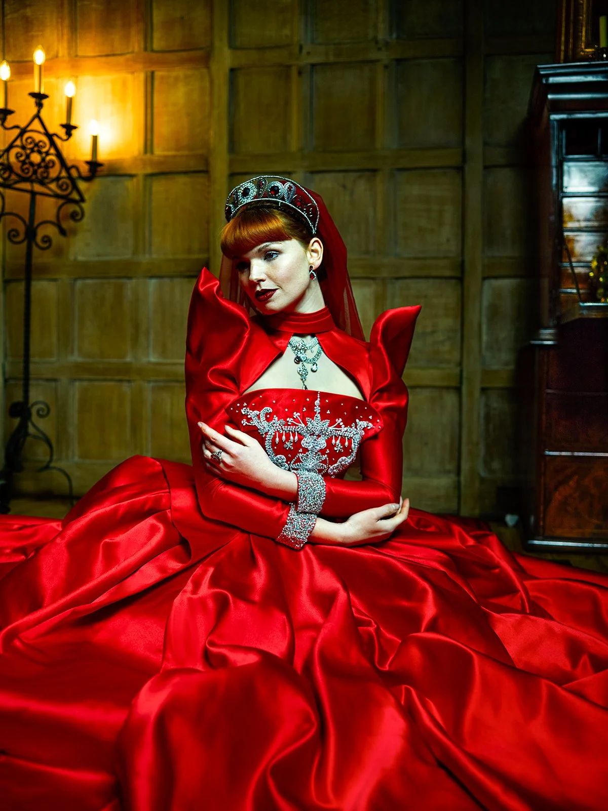 A woman in a red gown and crown, sitting on the floor with a wooden paneled wall behind her, decorated with a chandelier and some furniture.