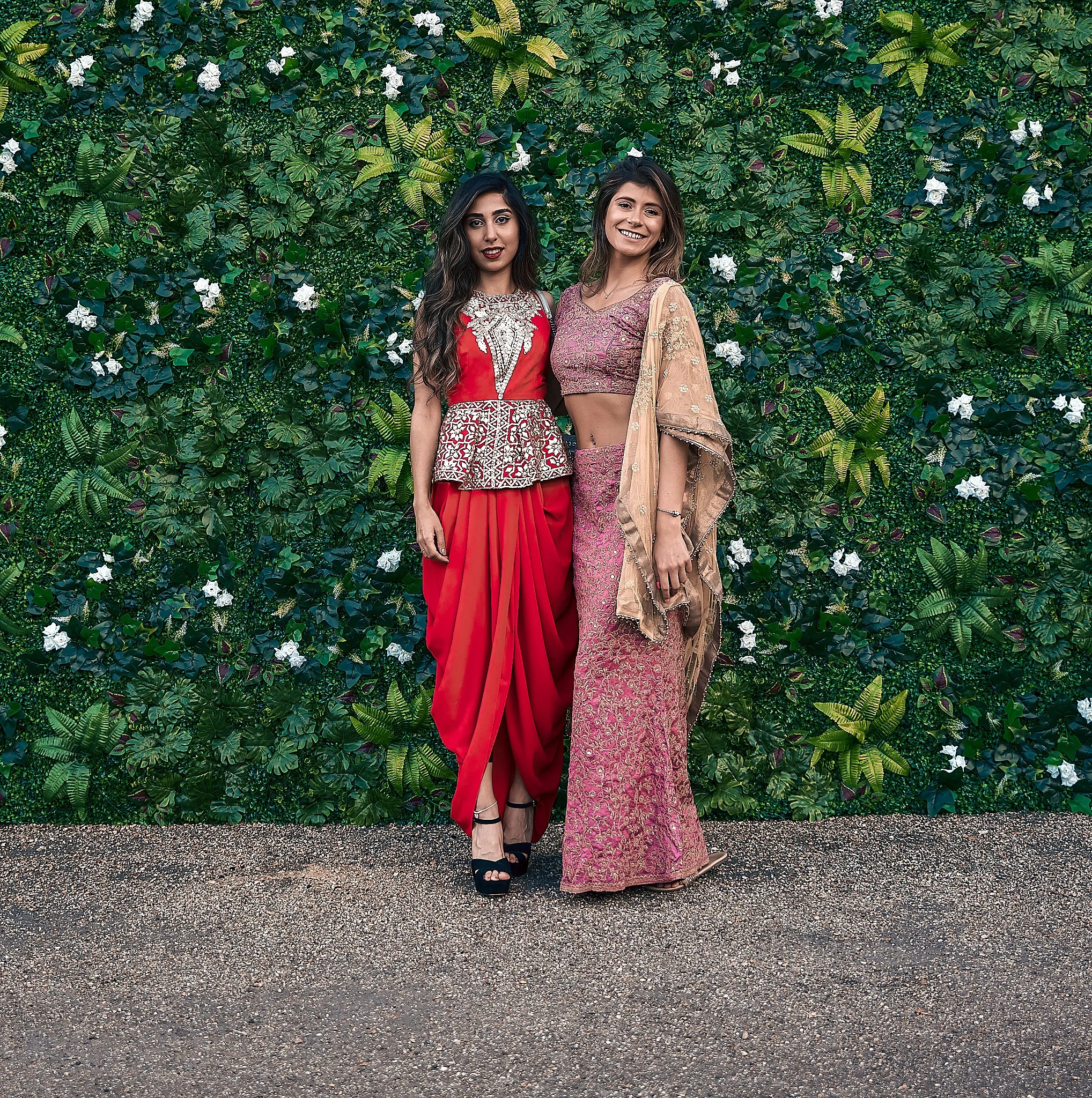 Two women in traditional Indian attire standing in front of a lush green leafy wall decorated with white flowers, smiling at the camera.