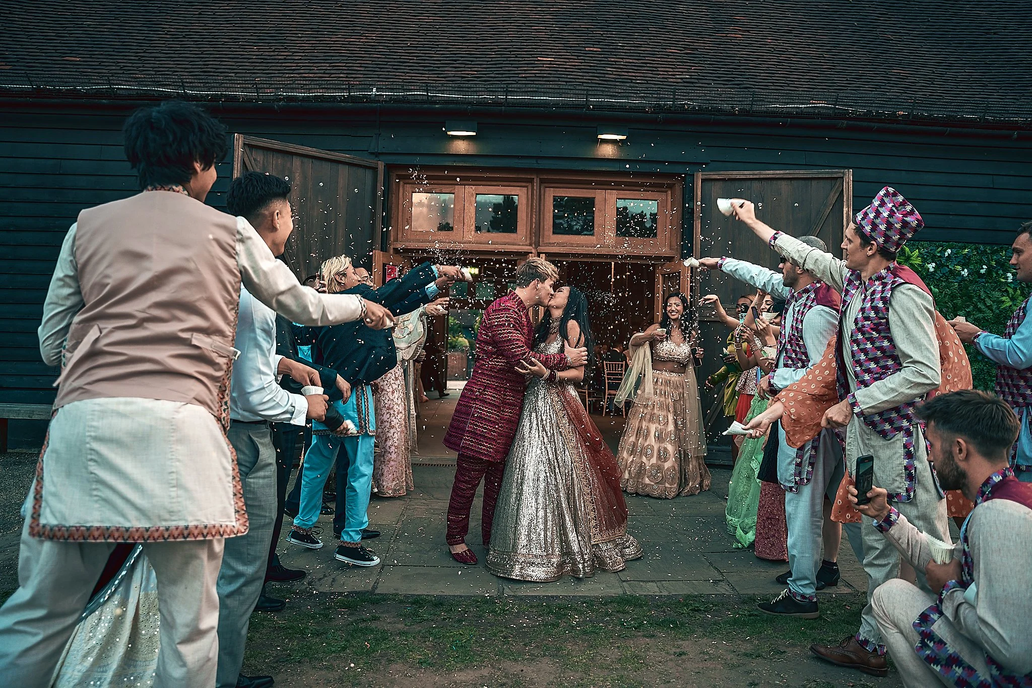 Couple in traditional Indian attire kissing as friends celebrate with water and flower toss outside of a house.