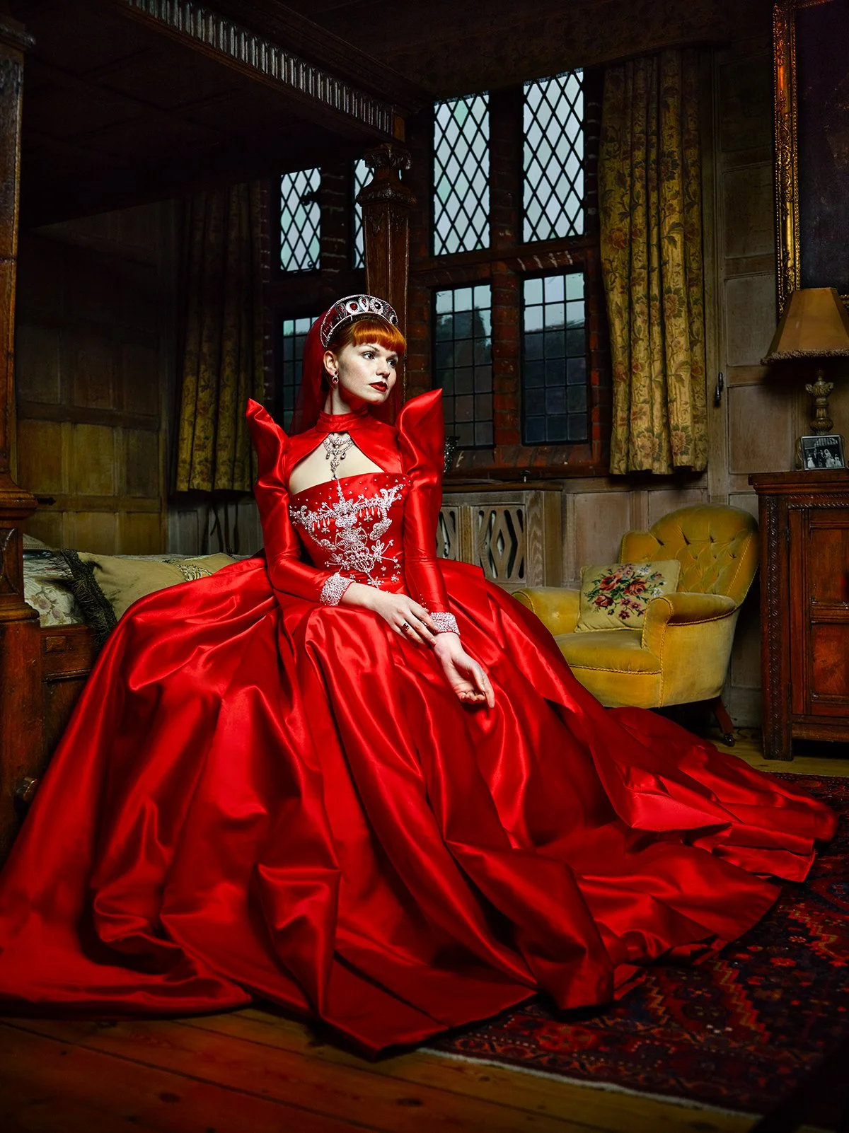 A woman wearing an elegant, red, Victorian-style gown with intricate white embroidery, sitting on a wooden bed in a vintage room with wood-paneled walls, large multi-paned windows, yellow armchair, and floral curtains.