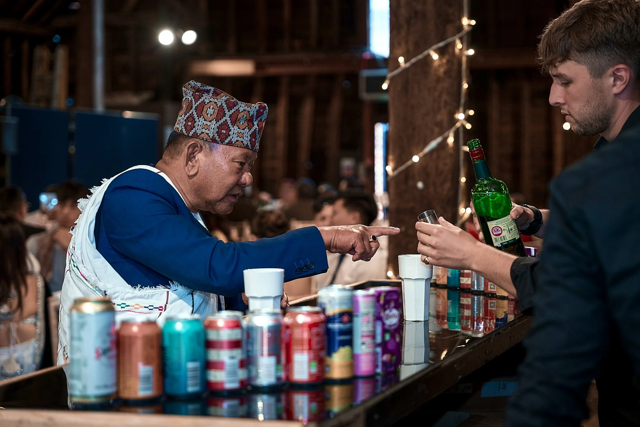 A bartender pouring a drink from a green bottle into a shot glass for a customer at a bar with canned beverages on the counter, and a person pointing at him.