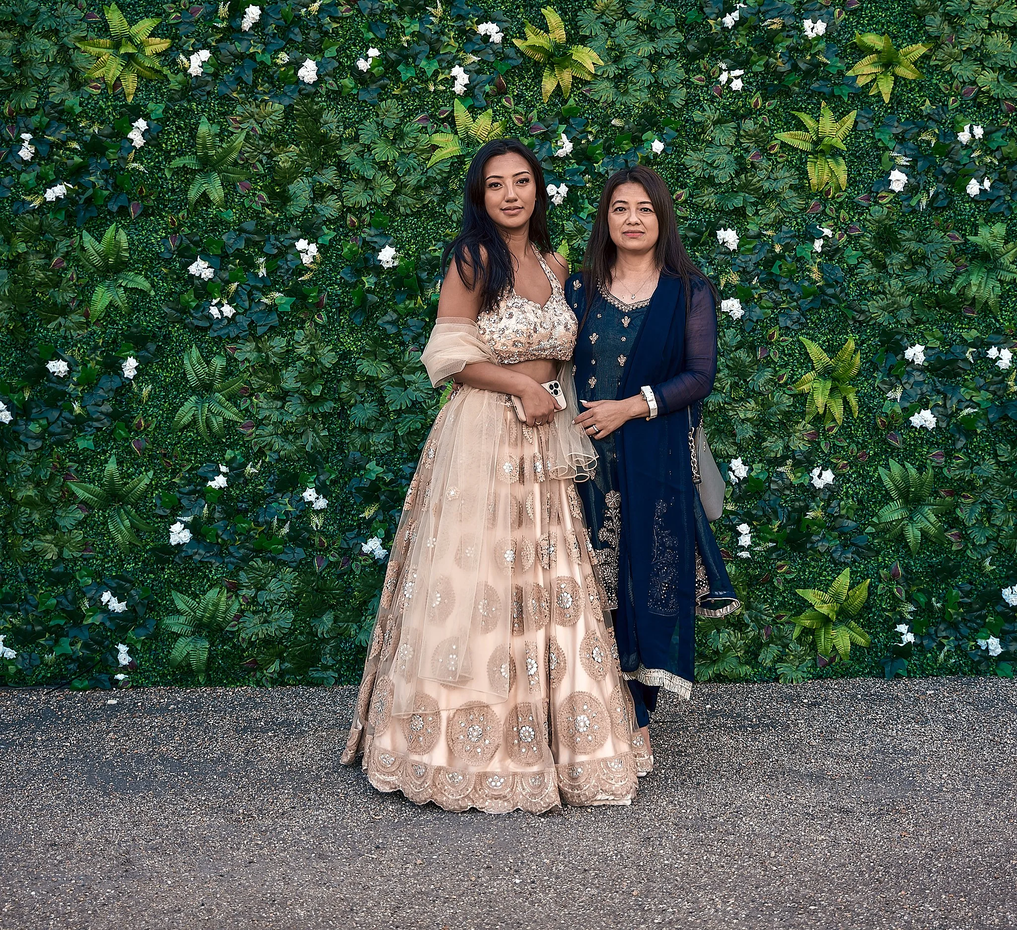 Two women standing in front of a leafy green wall with white flowers. The woman on the left is wearing a beige, embroidered gown with a sequined top and holding a phone. The woman on the right is wearing a dark navy traditional dress with gold embroi