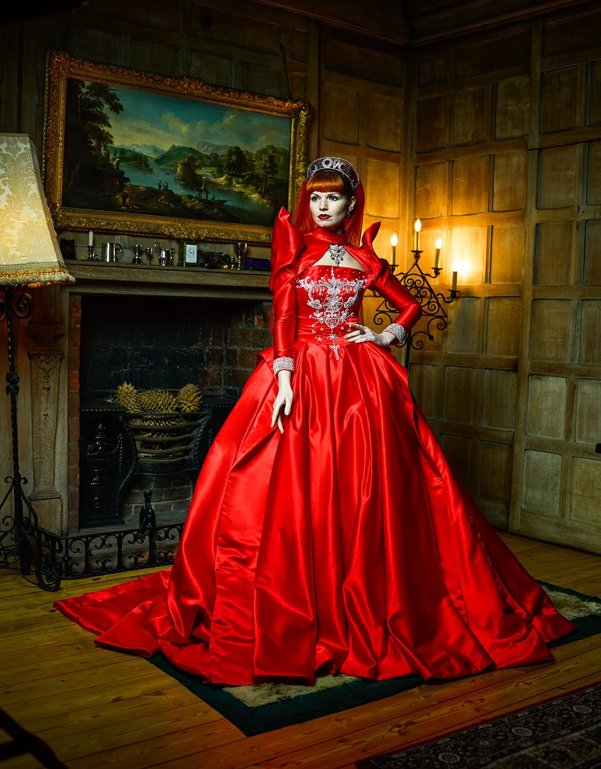 A woman in a luxurious red gown with intricate silver embroidery stands in a wood-paneled room with a fireplace and painting. She wears a silver crown or tiara and has red hair, striking makeup, and posed confidently.