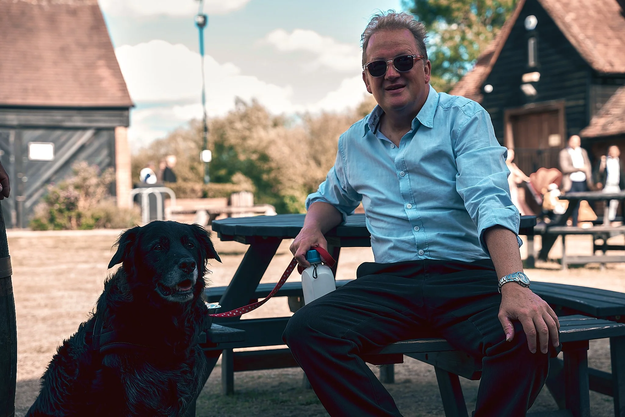 A man with sunglasses sits on a bench holding a leash attached to a black dog. The setting appears to be an outdoor park or café area with other people in the background and buildings with wooden exteriors.