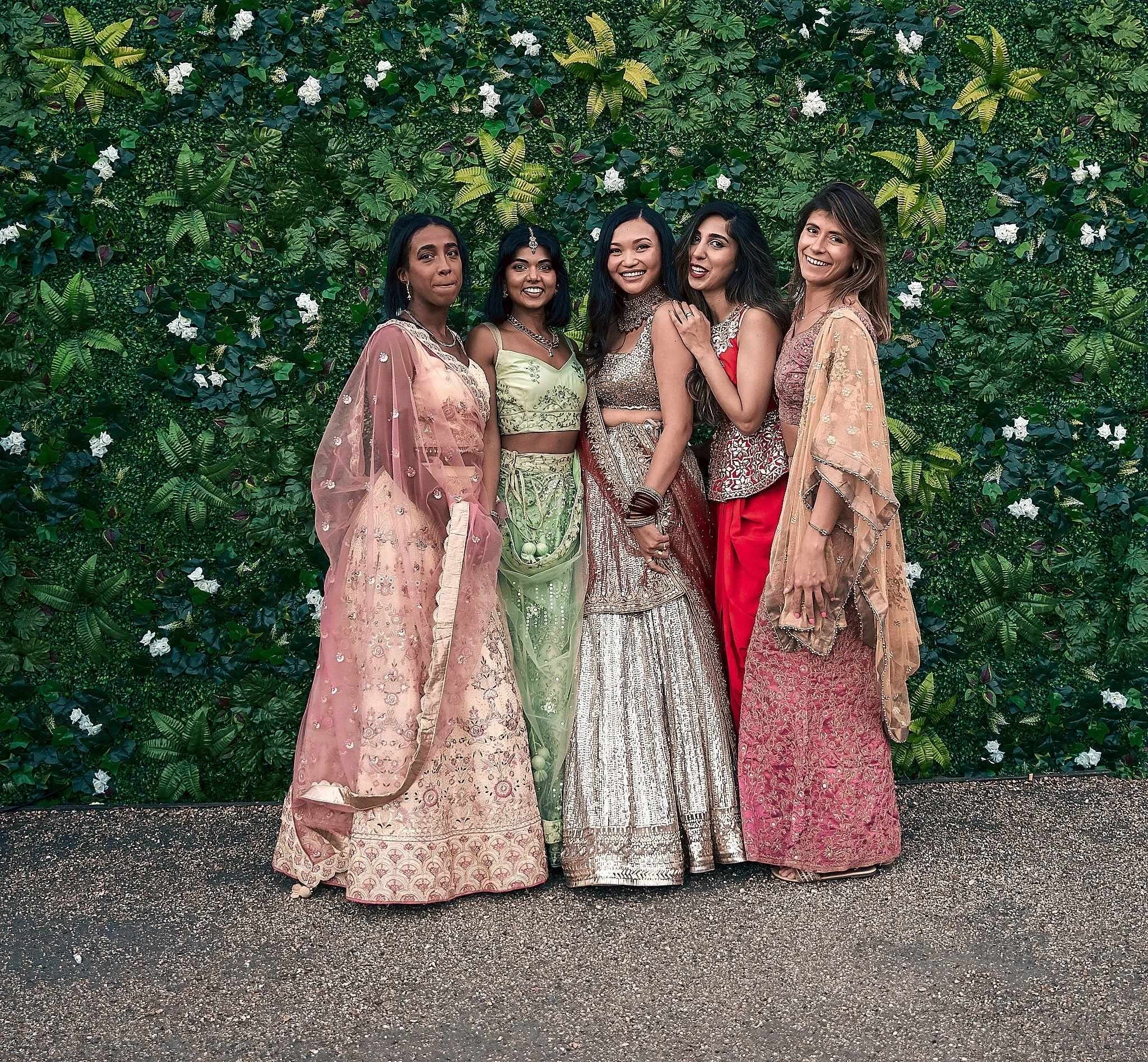 Five women in colorful traditional Indian attire pose in front of a lush green floral backdrop, smiling and standing close together.