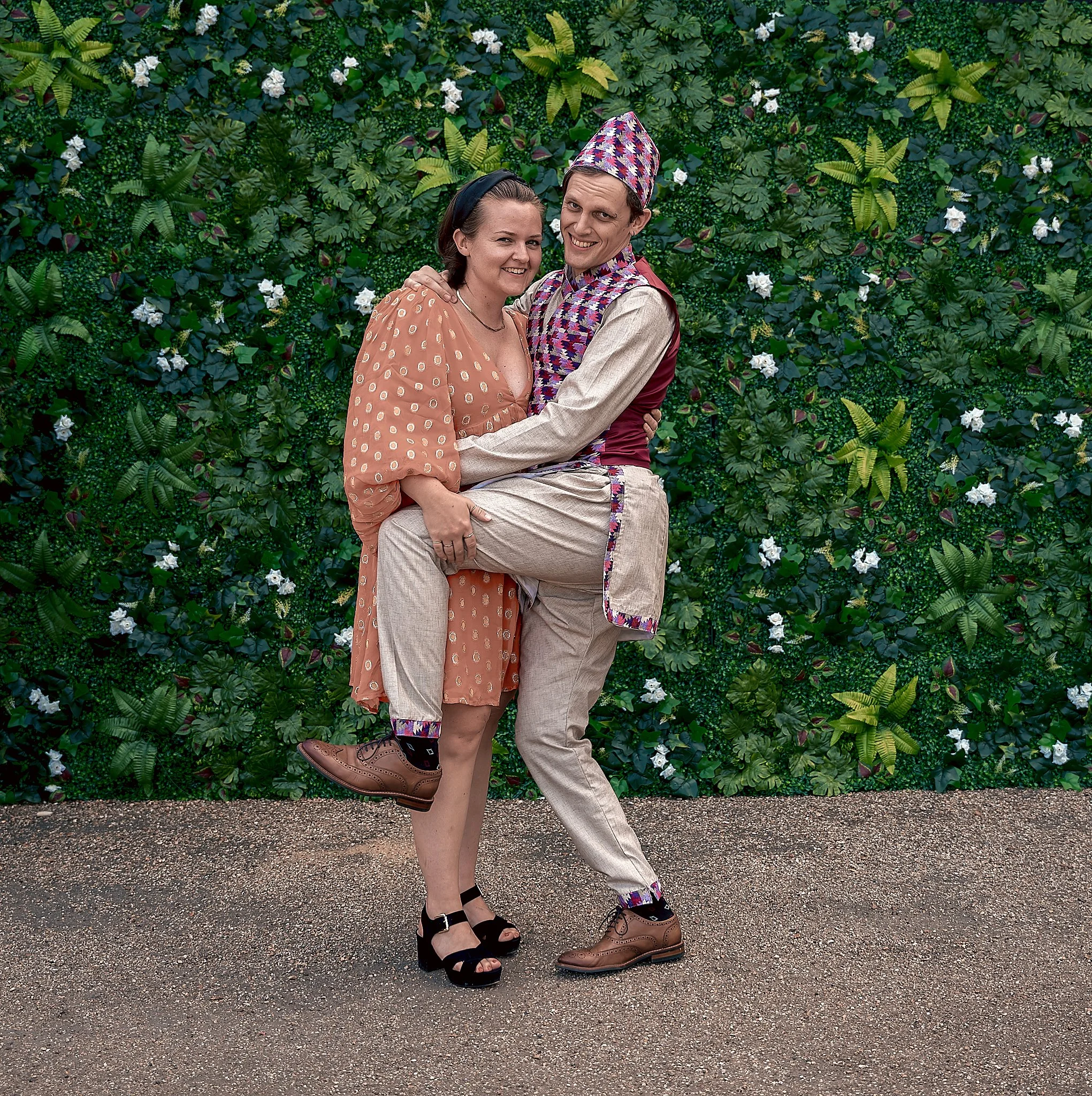 Two people dressed in colorful, vintage-style clothing, posing in front of a lush green floral wall.