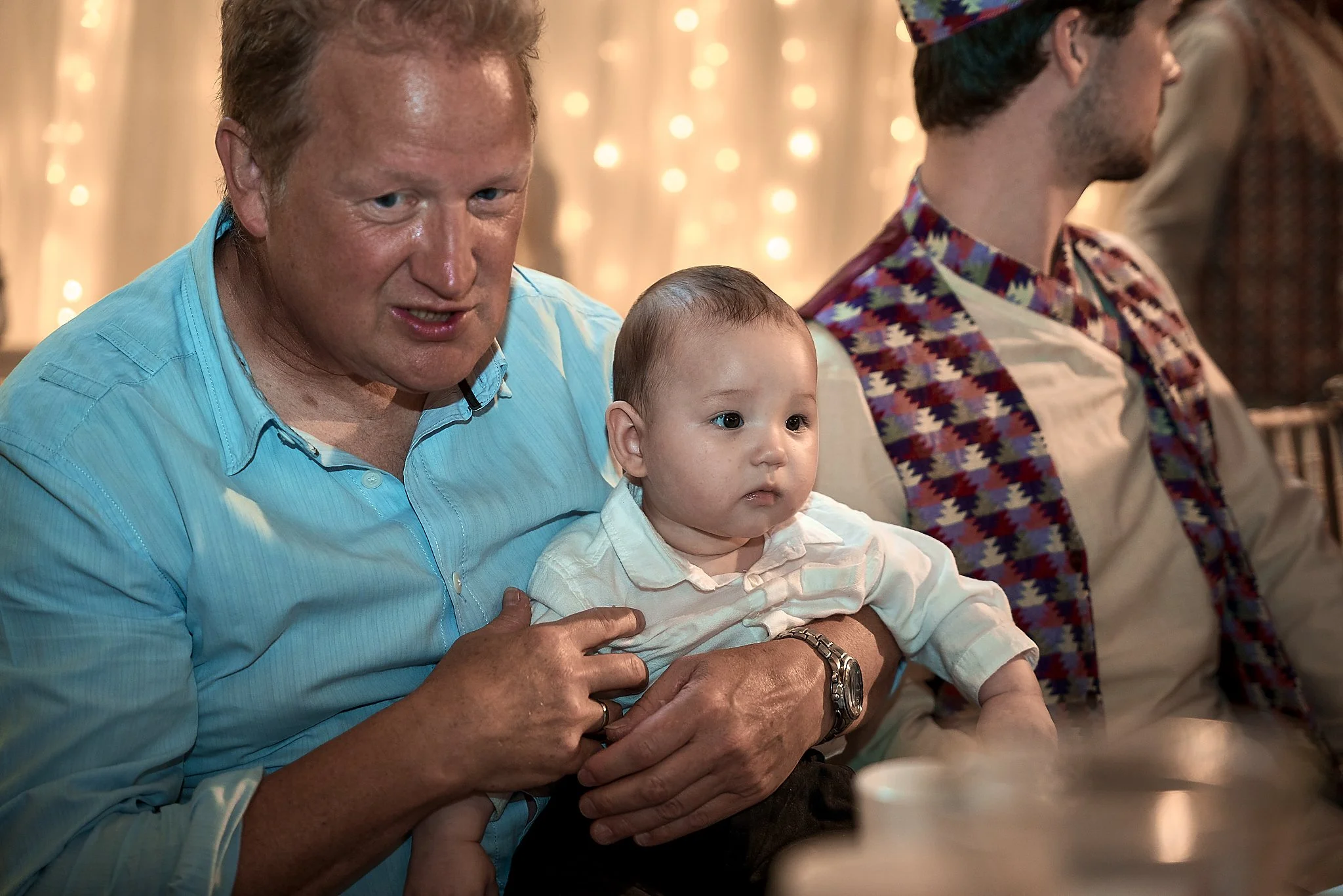 A man holding a young child at a social gathering. The man has red hair and is wearing a light blue shirt, while the child is wearing a white shirt. There are other people in the background, and the setting appears to be festive with warm lighting.