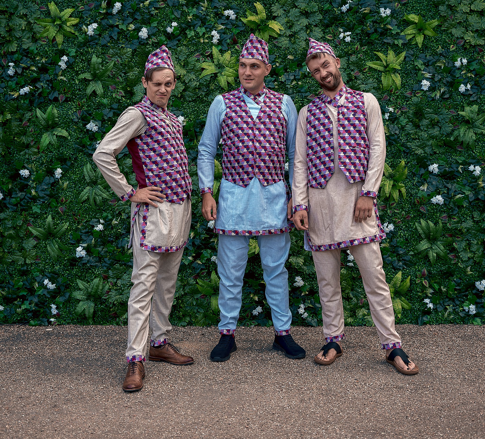 Three men dressed in colorful, patterned traditional clothing standing in front of a lush green wall with white flowers.