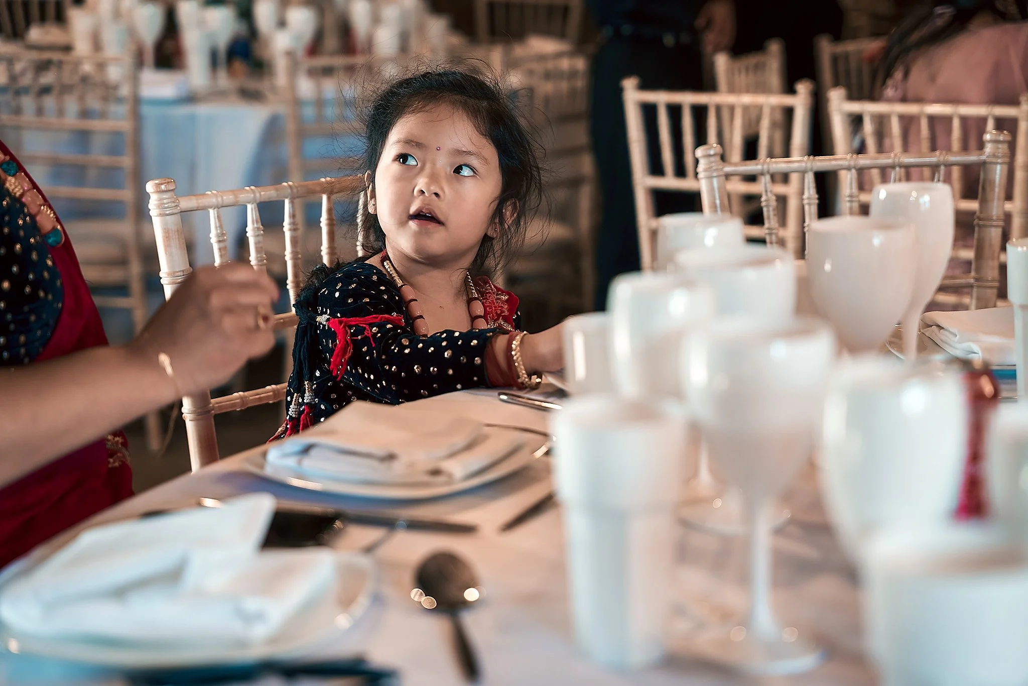 A young girl with black hair and traditional clothing sitting at a banquet table, looking to her right, surrounded by white tableware and chairs in a decorated event hall.