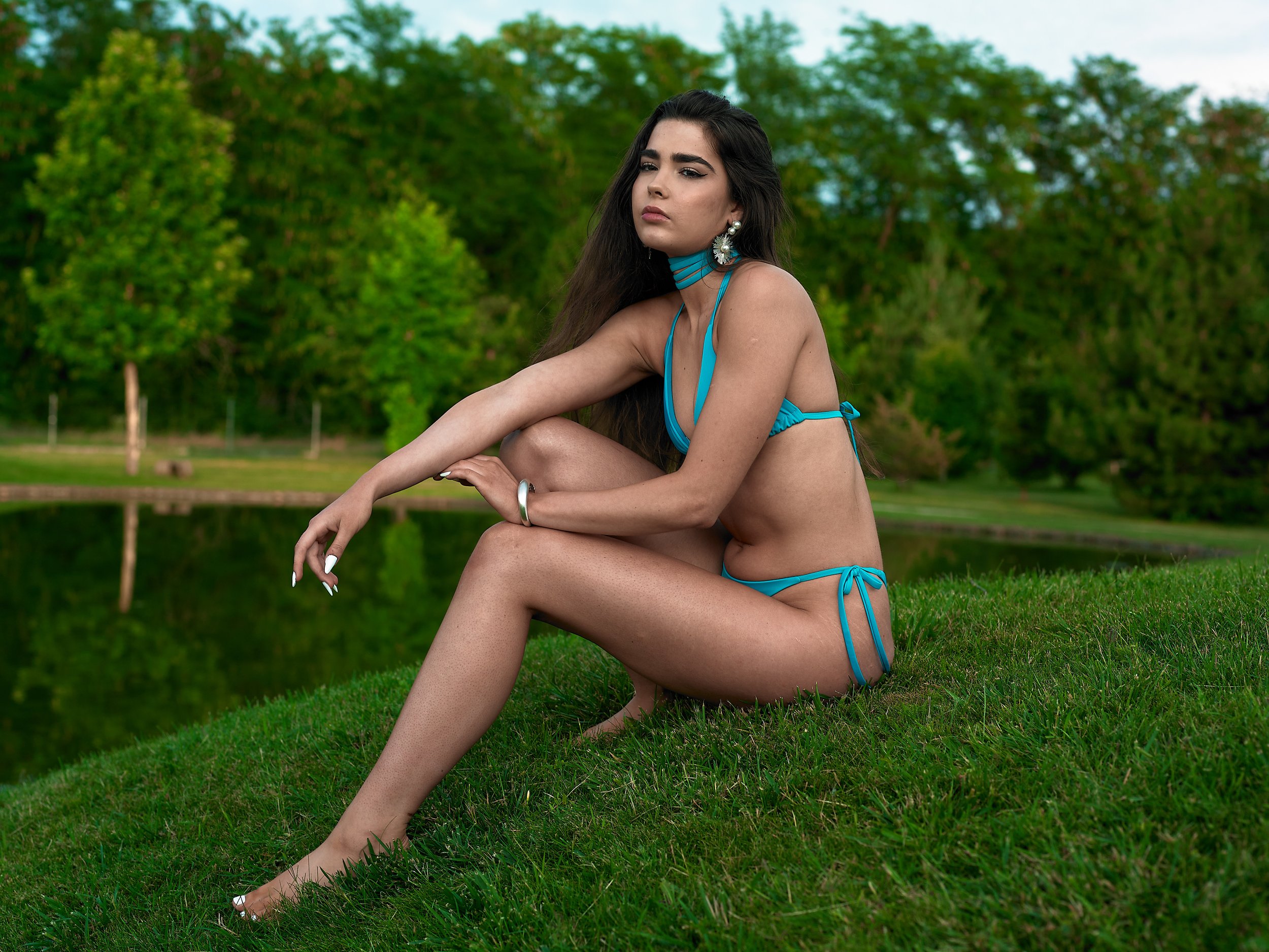 A woman with long dark hair, wearing a blue bikini, sitting on grass near a body of water in a park with green trees in the background.