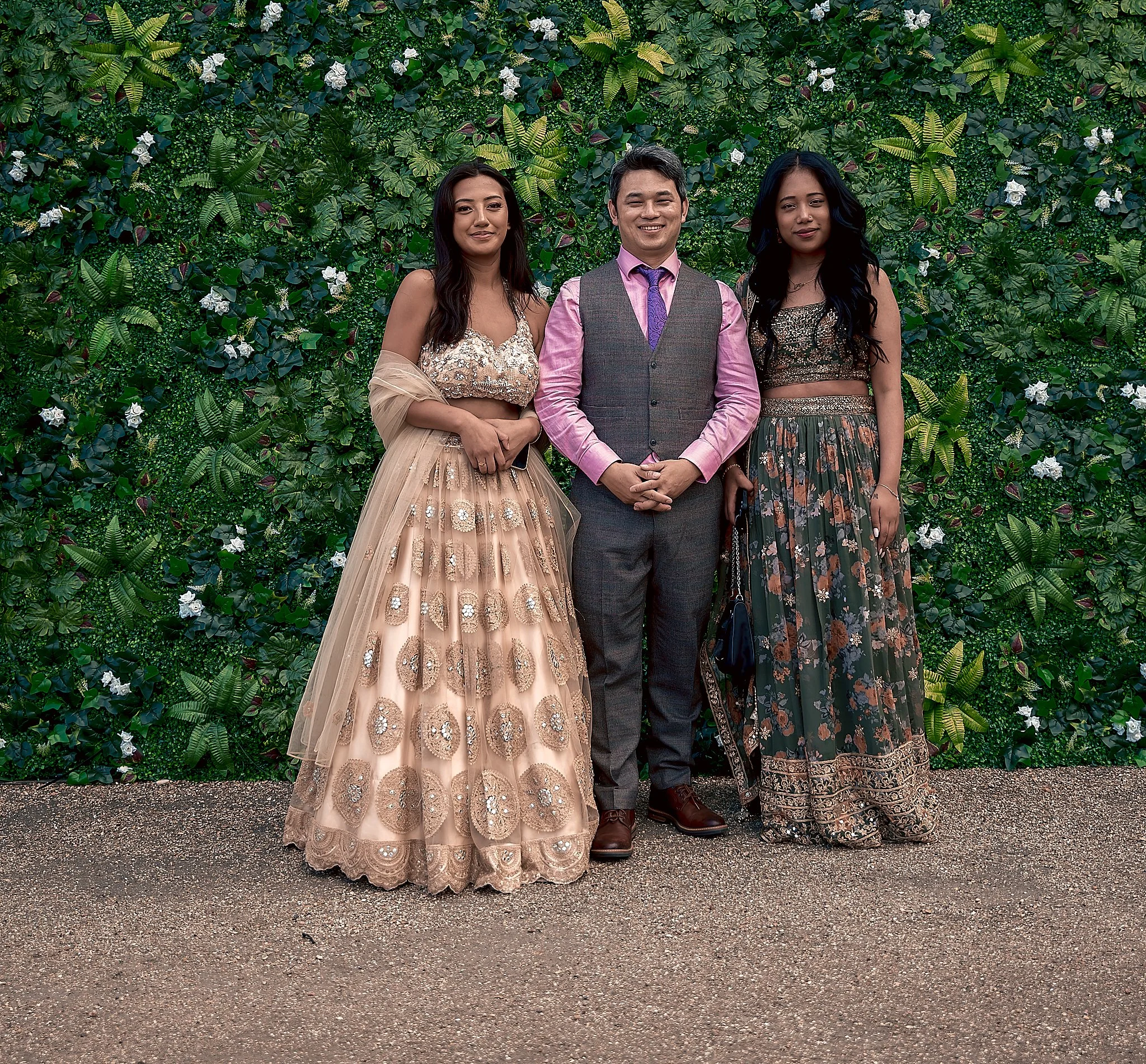 Three people standing in front of a green leafy wall. The woman on the left is wearing a beige embellished gown with a beige shawl. The man in the middle is dressed in a pink shirt, gray vest, and matching gray trousers. The woman on the right is wea
