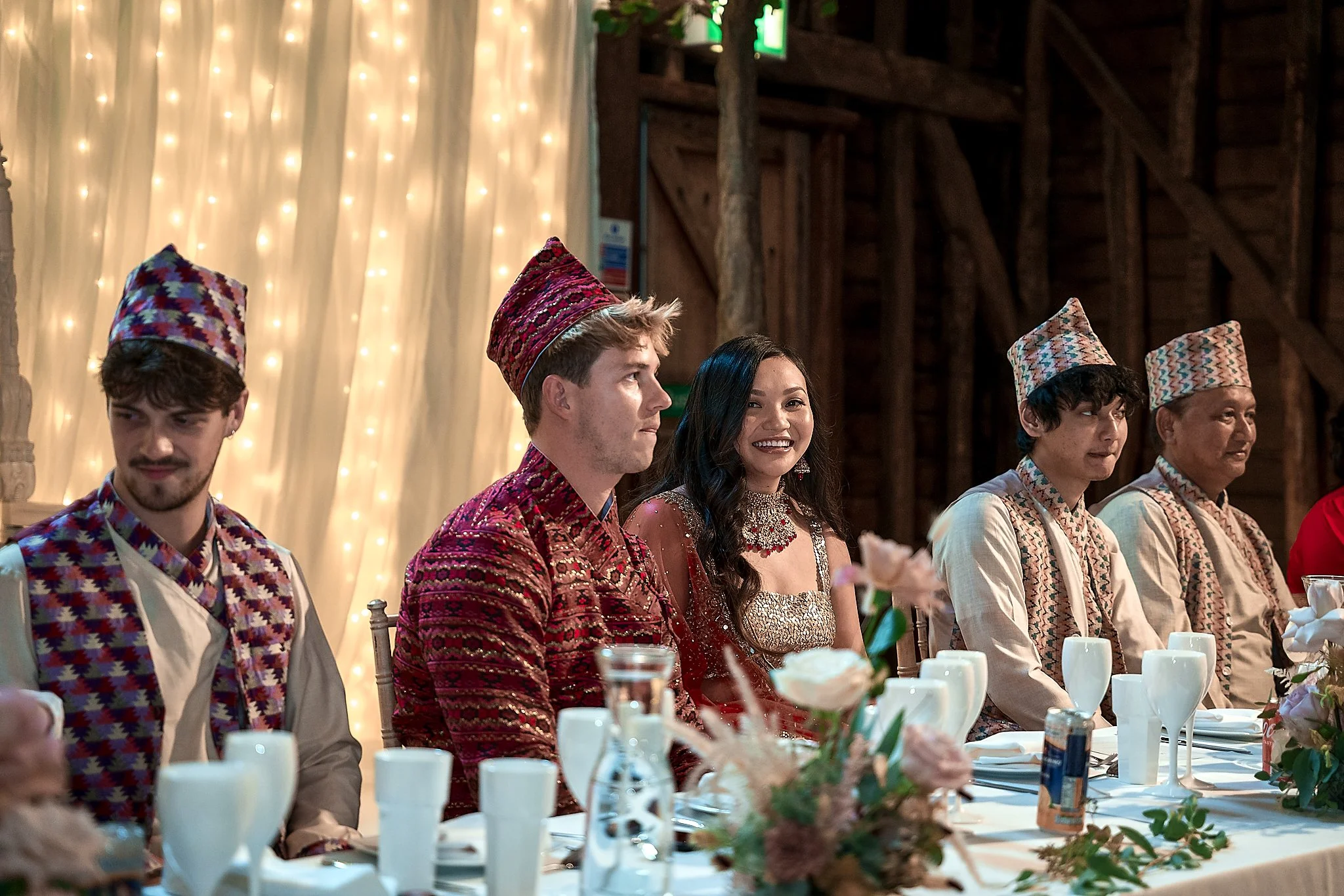 People dressed in traditional Nepali attire, sitting at a decorated table during a celebration or wedding, with a backdrop of fairy lights.