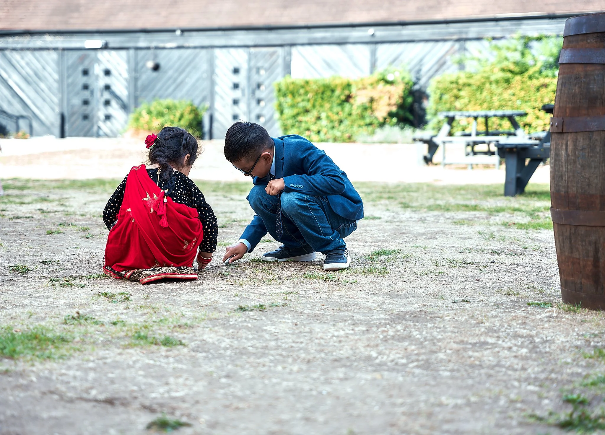 Two children, a girl and a boy, crouching on the ground and exploring something in an outdoor area with dirt and sparse grass, with a wooden fence and trees in the background.