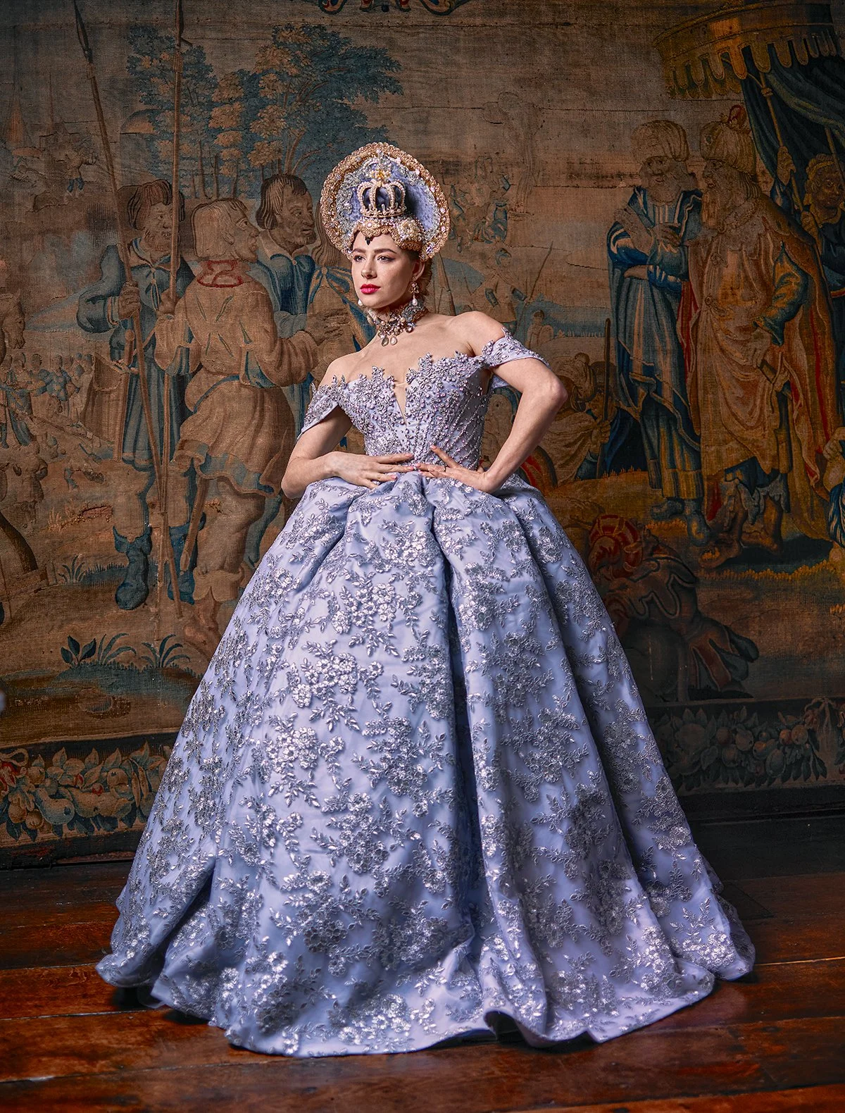 A woman in a regal, off-the-shoulder lavender ball gown with silver embroidery and lace, wearing a large ornate crown and jewelry, standing with one hand on her waist against a historical tapestry backdrop.