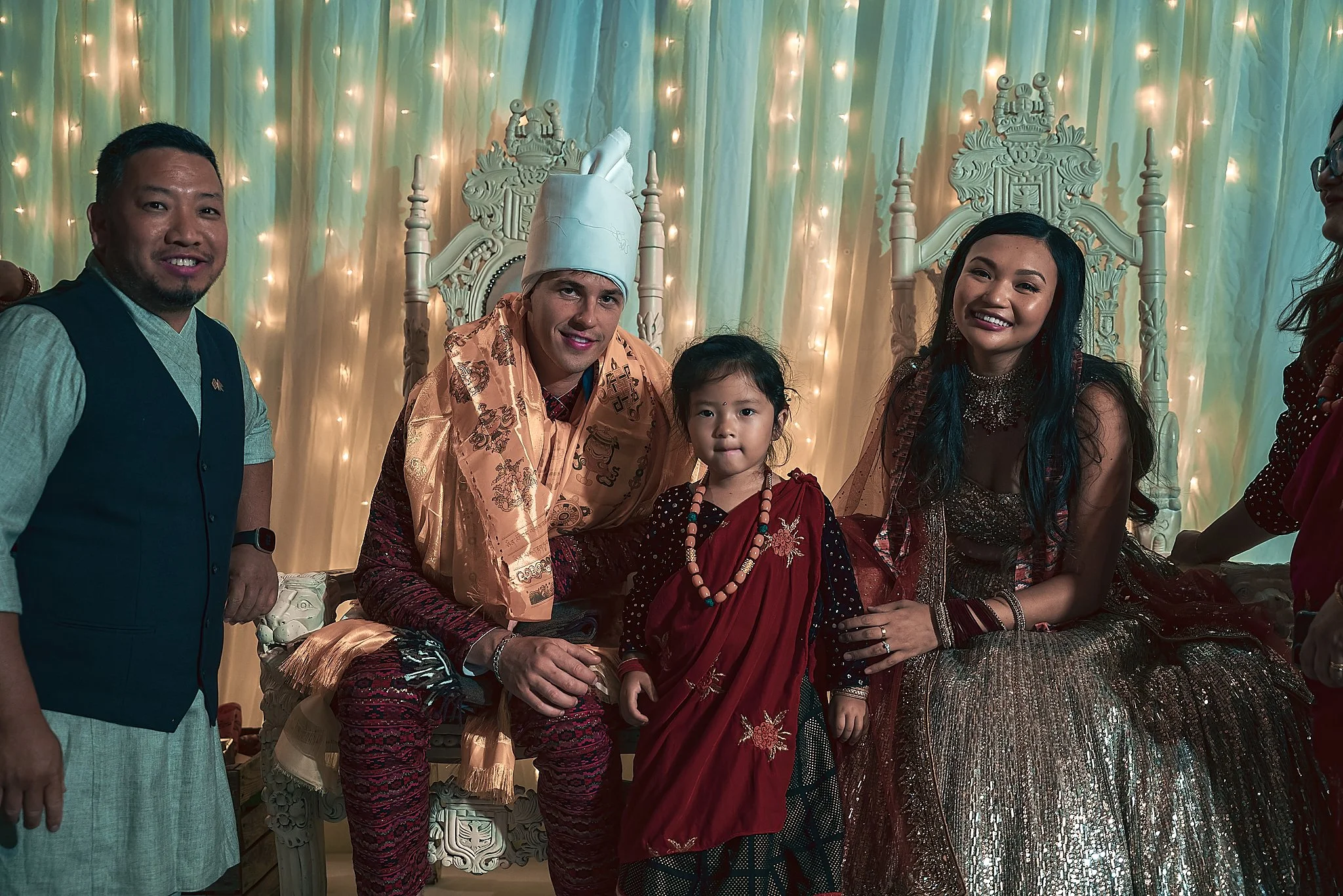 People celebrating a special event, with a girl in traditional attire standing between a seated man in ceremonial clothing and a woman in an elegant dress, in front of a decorated backdrop with lights.
