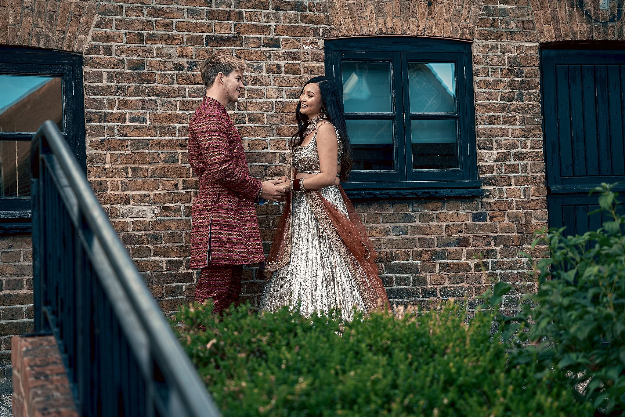A man and woman dressed in traditional Indian attire facing each other, holding hands, standing outdoors against a brick wall with blue window frames.