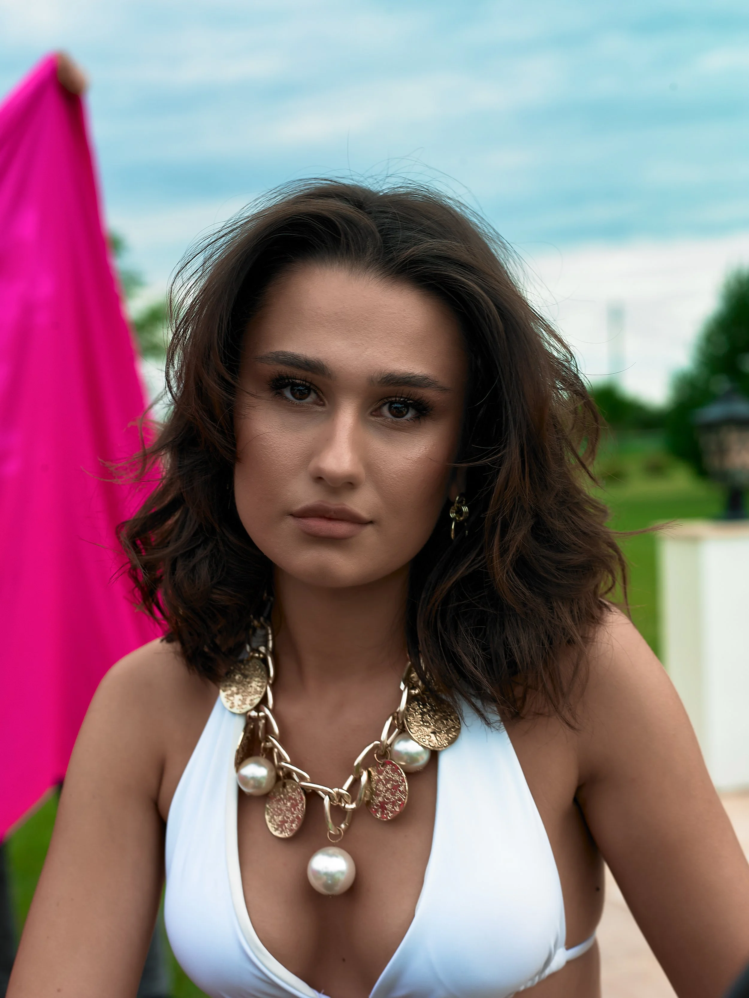 A young woman with dark wavy hair, wearing a white top and gold jewelry, sitting outdoors with a pink umbrella and cloudy sky in the background.